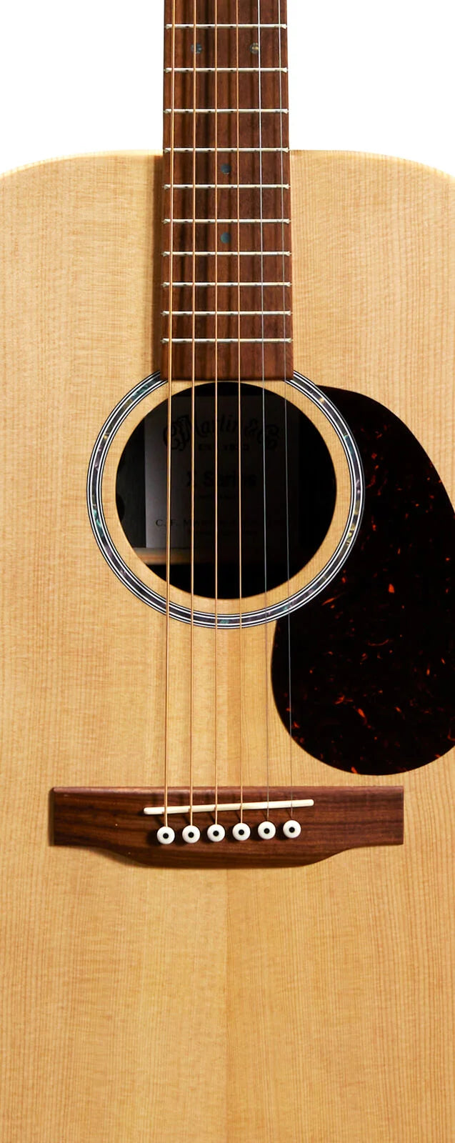 Natural wood acoustic guitar body with six strings, a round soundhole bordered by rings, dark pickguard, and a wooden bridge with white bridge pins.