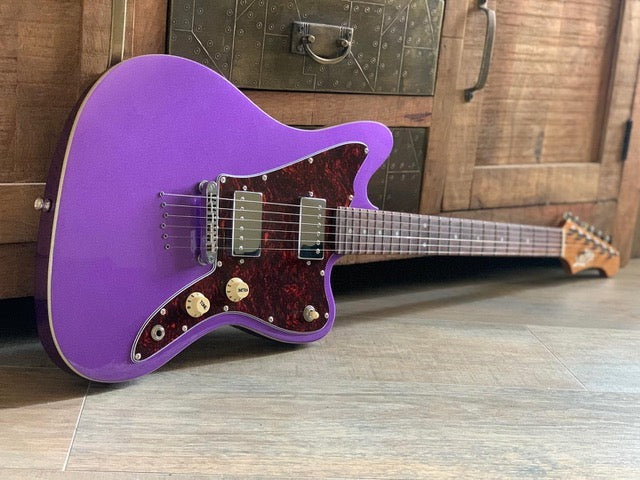 Purple electric guitar with a dark reddish-brown pickguard, dual humbucker pickups, three control knobs, and a wooden fretboard, resting on a wood floor.