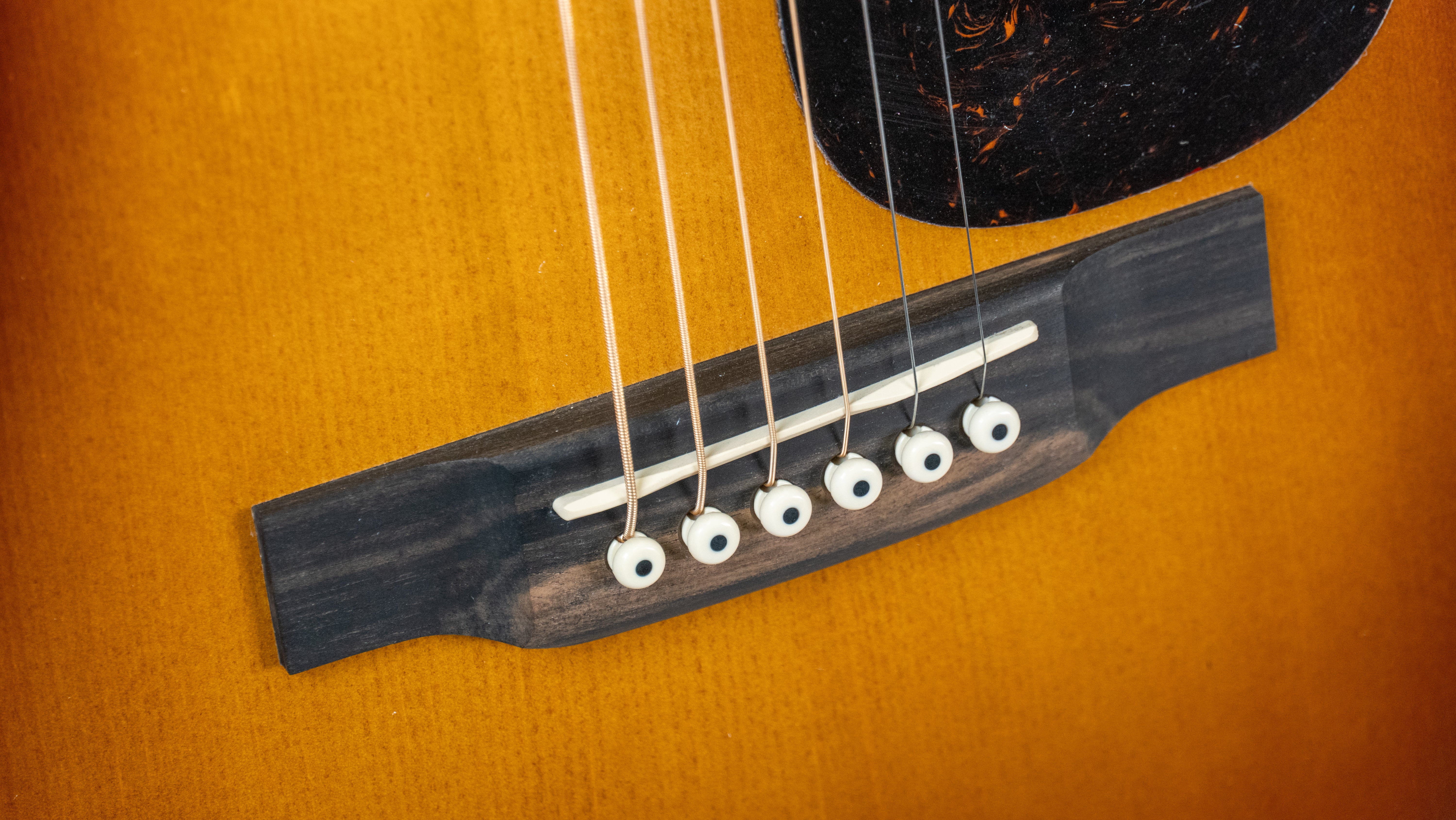 Close-up of an acoustic guitar bridge showing six strings anchored by white bridge pins on a warm brown wood body.