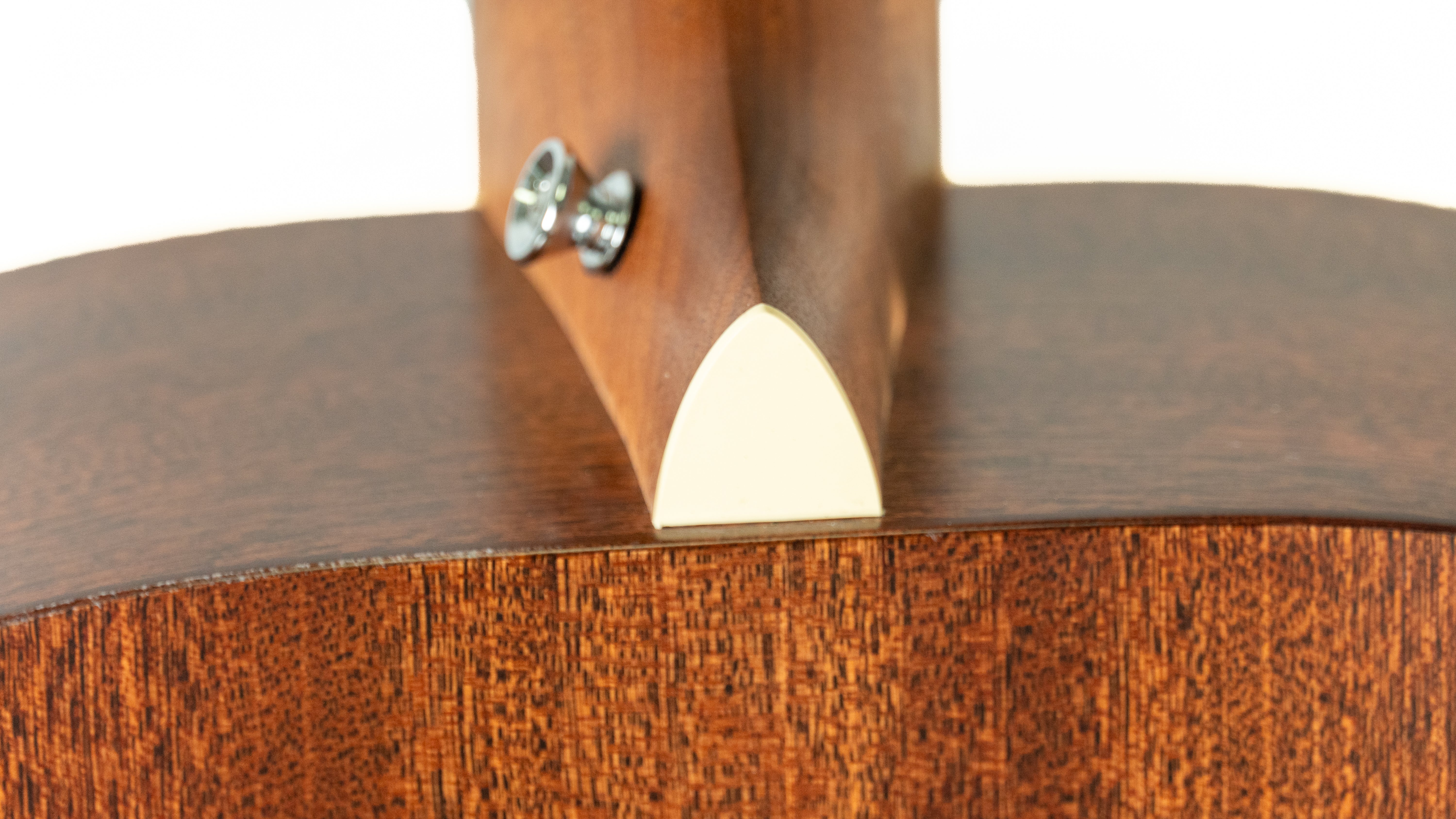 Close-up of an acoustic guitar’s neck joint, showing glossy brown wood, a cream triangular heel cap, and two shiny metal strap buttons.
