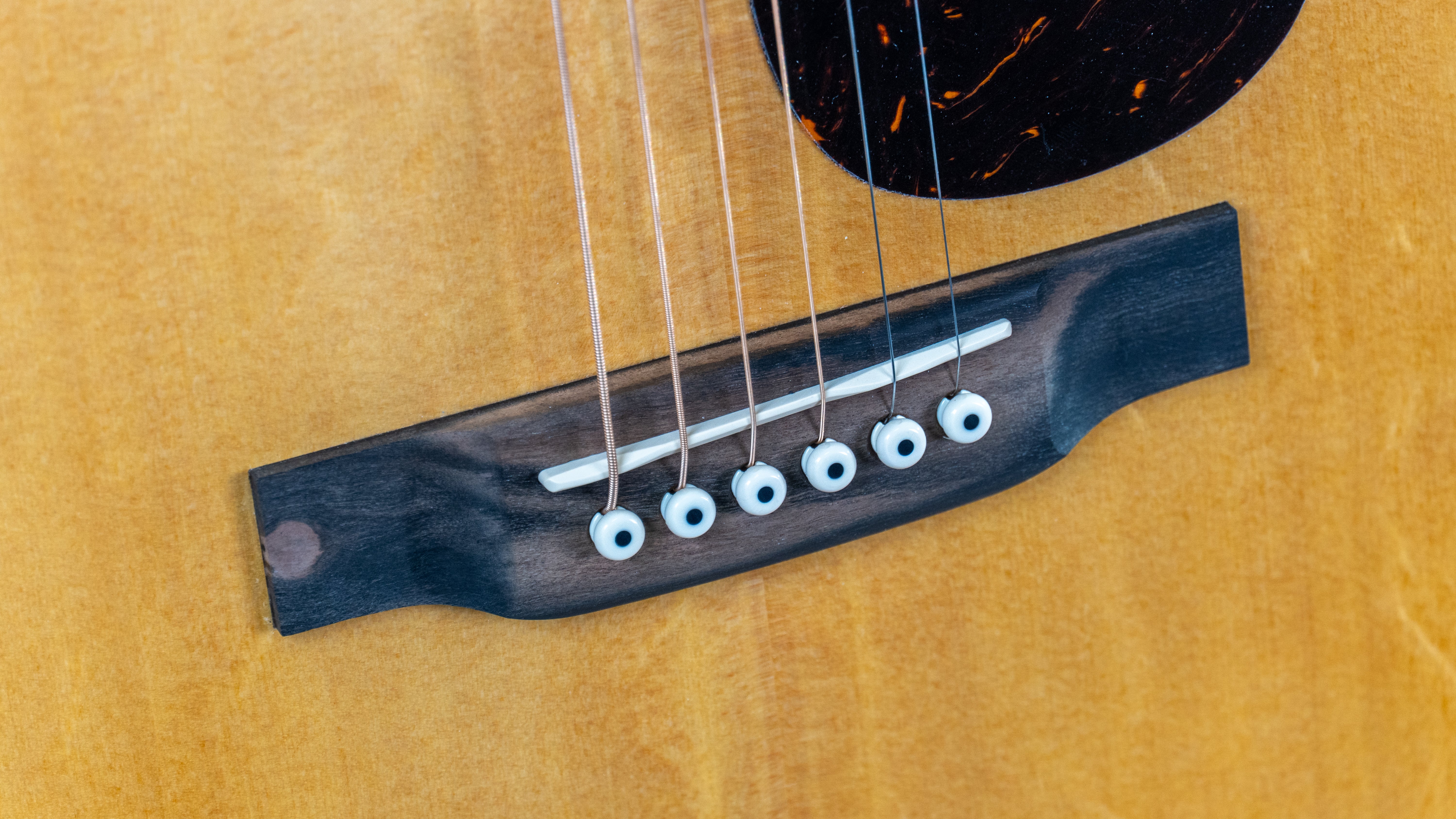 Close-up of an acoustic guitar bridge showing six strings, white bridge pins, and part of a black pickguard on a light wood body.