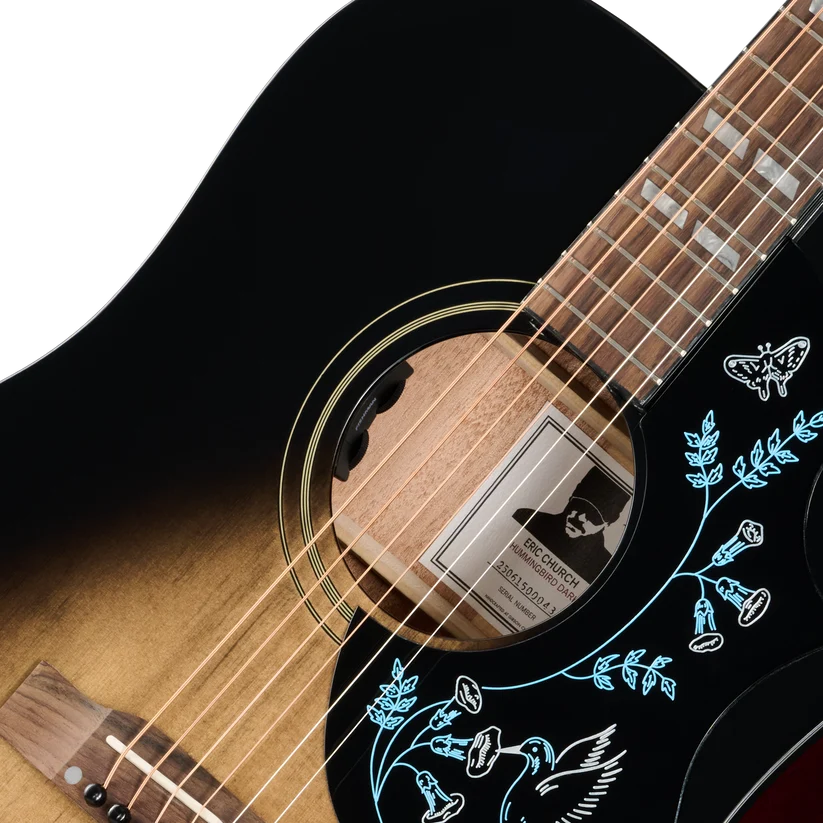 Acoustic guitar with a dark sunburst body, floral and bird designs around the sound hole, and a label featuring a silhouette of a person inside.