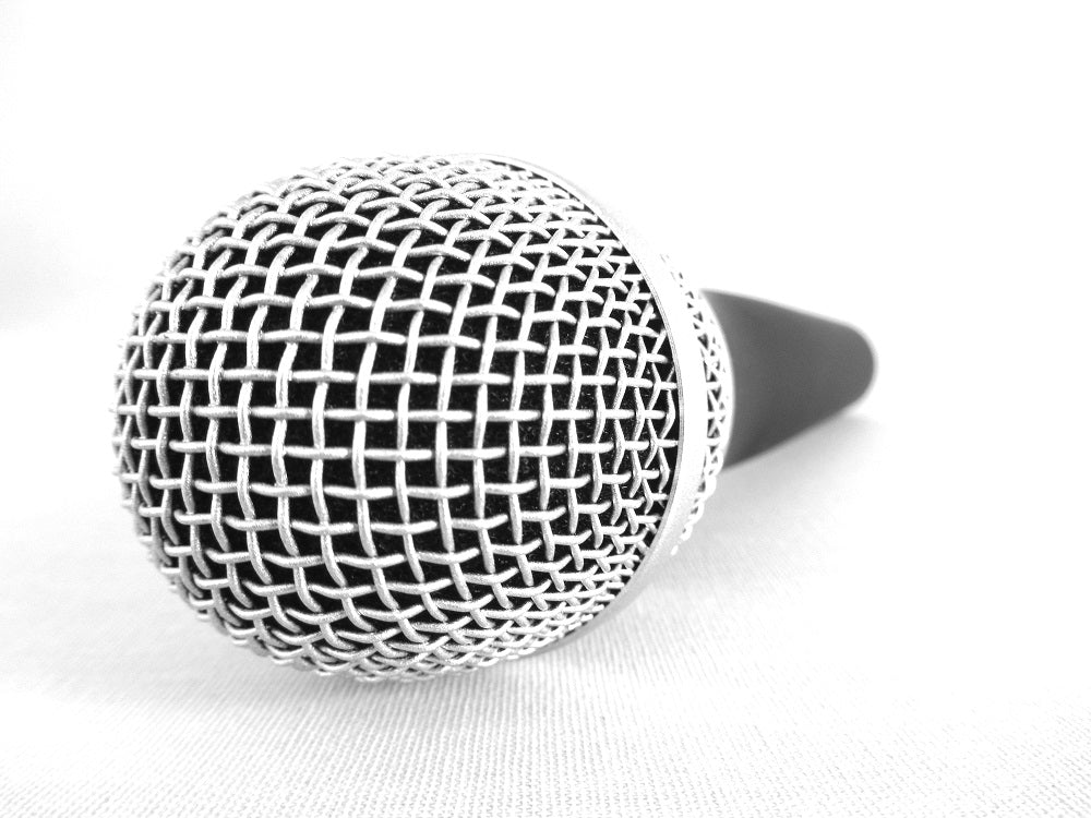 A handheld recording microphone with a black handle and silver mesh grille rests on a white surface, viewed close up from the front.