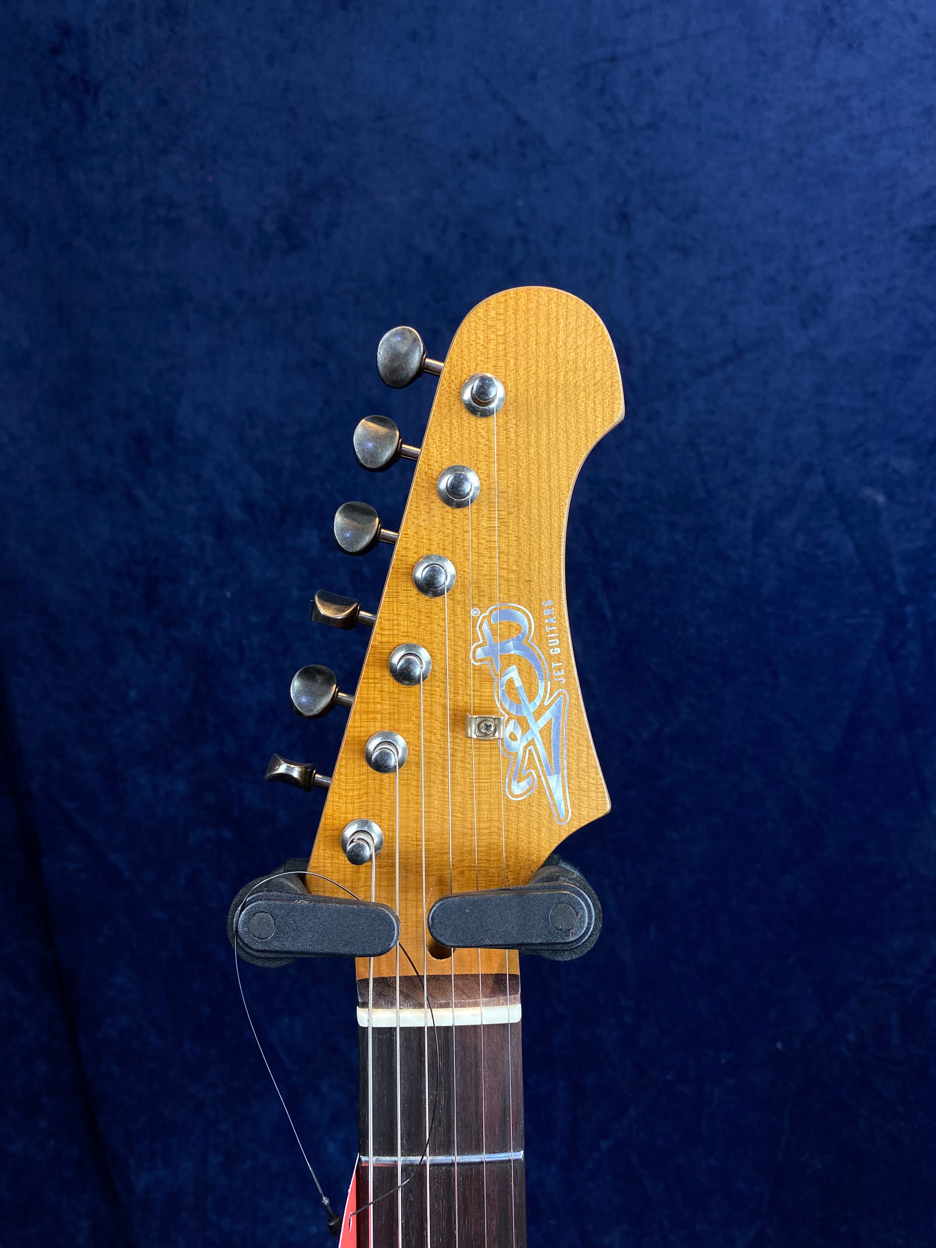 Light brown electric guitar headstock with six tuning pegs, metallic logo, and strings against a dark blue background.