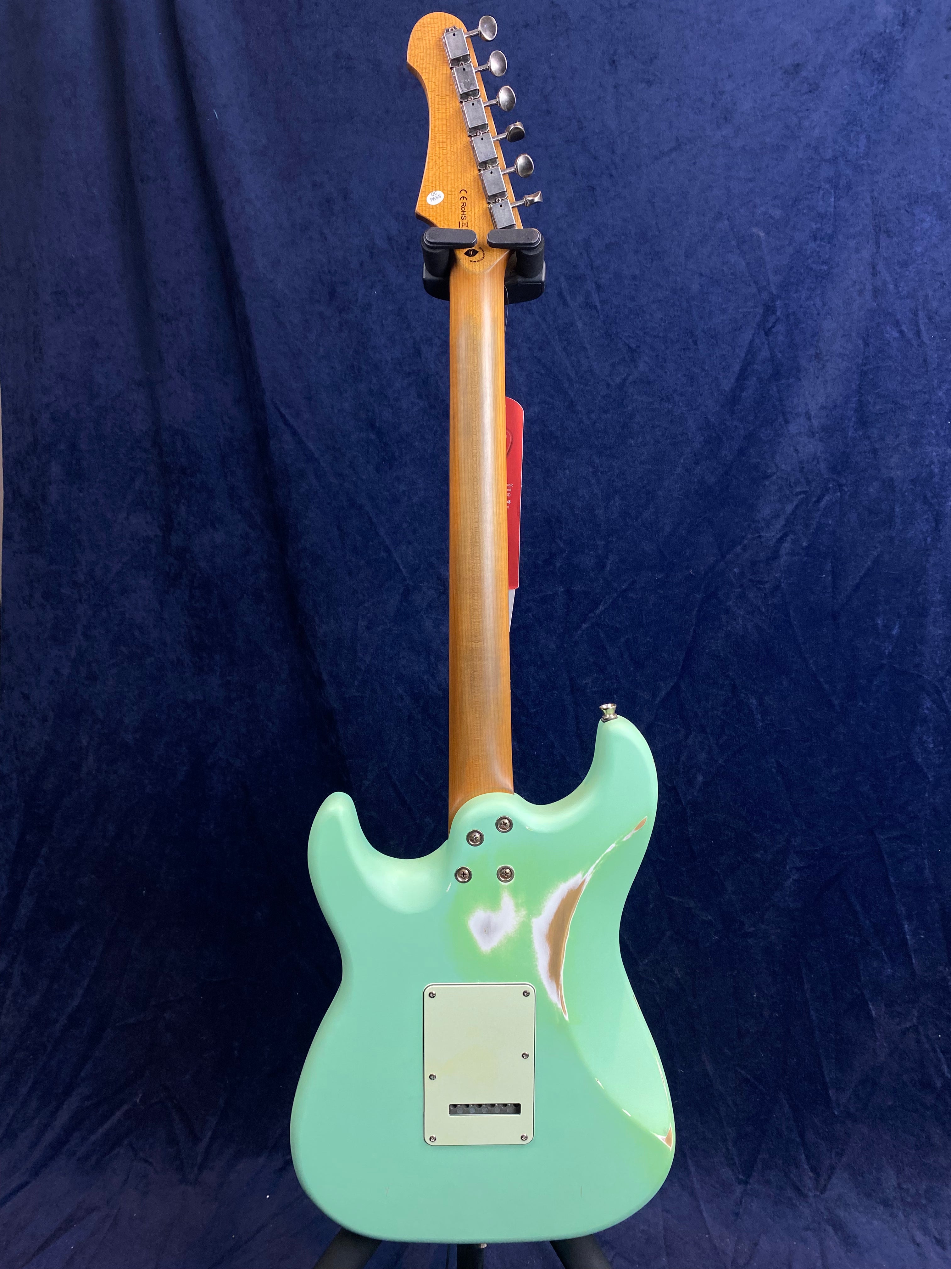 A green electric guitar with a worn finish revealing wood patches on the back, maple neck, and silver tuners, shown from the rear against a dark background.