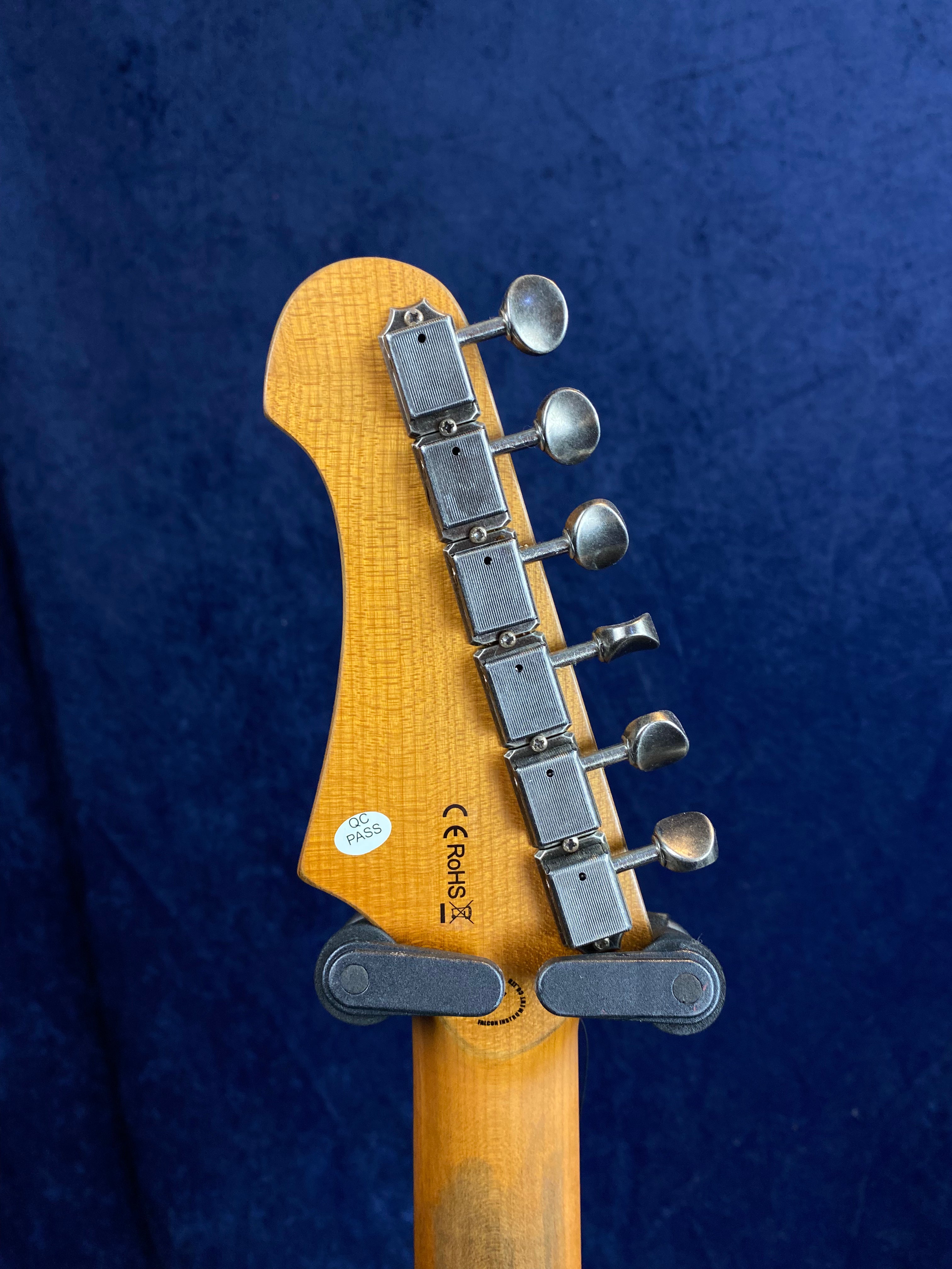 Back of an electric guitar headstock with six metal tuning pegs, wood grain visible, QC Pass sticker, and certification marks above the neck.