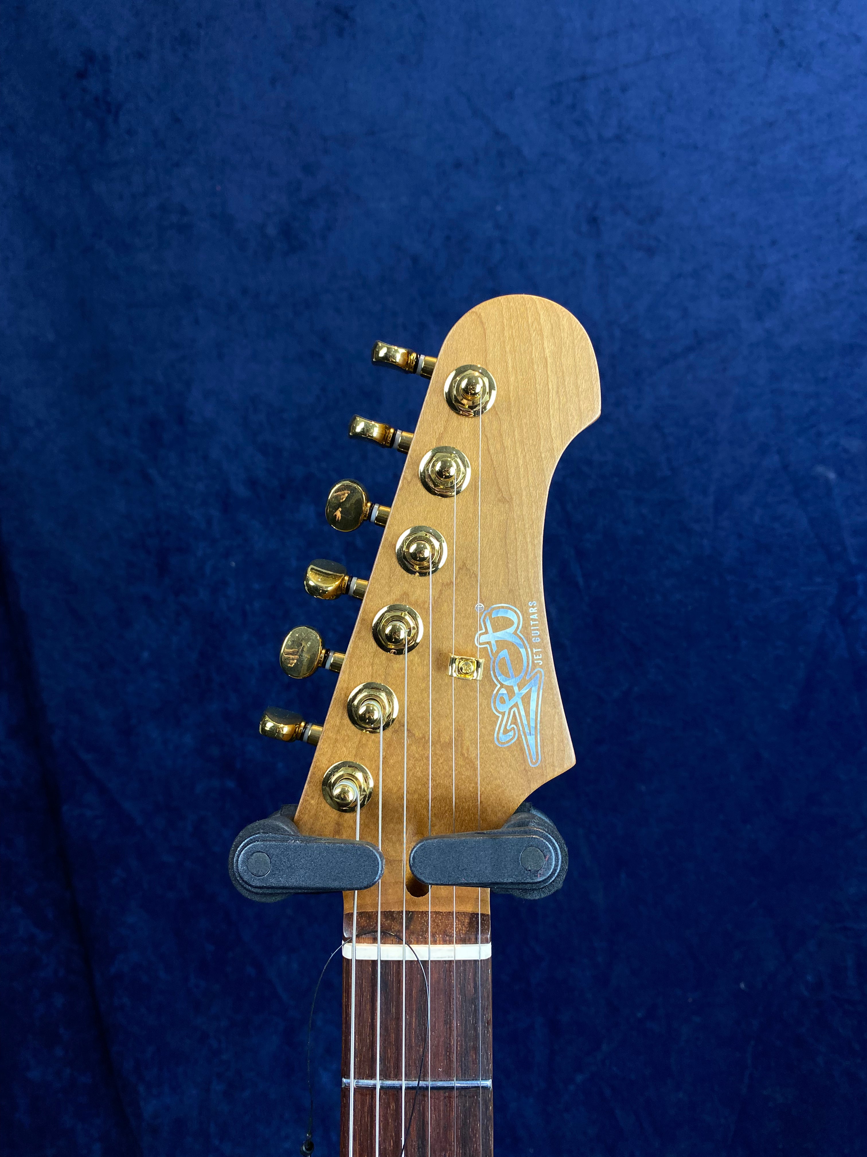 Jet Guitars headstock with six gold tuning pegs, light wood finish, and Jet logo on the front, set against a dark blue background.