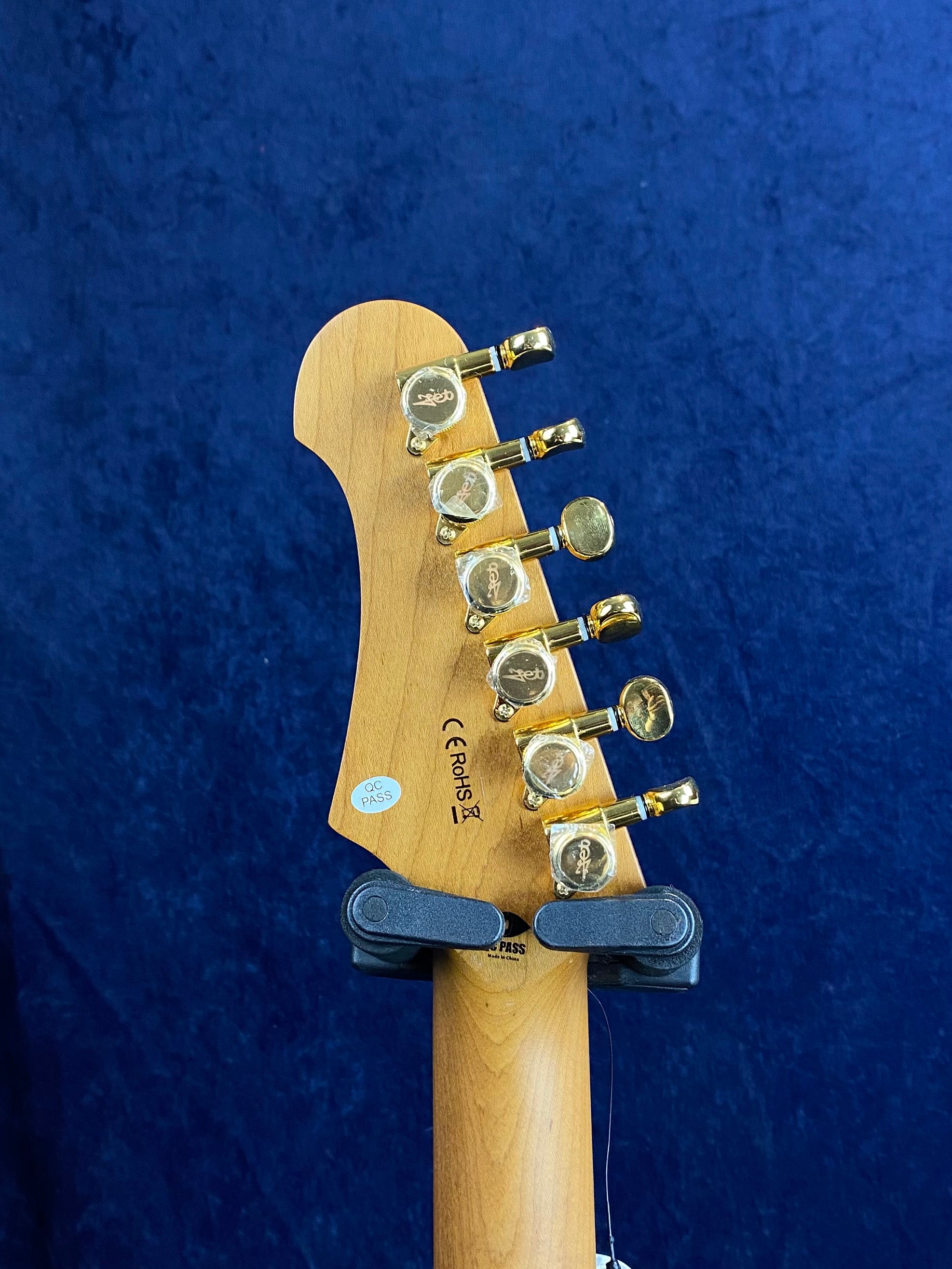 Jet Guitar headstock back view with six gold tuning pegs, maple wood finish, and certification stickers, held upright against a dark blue background.