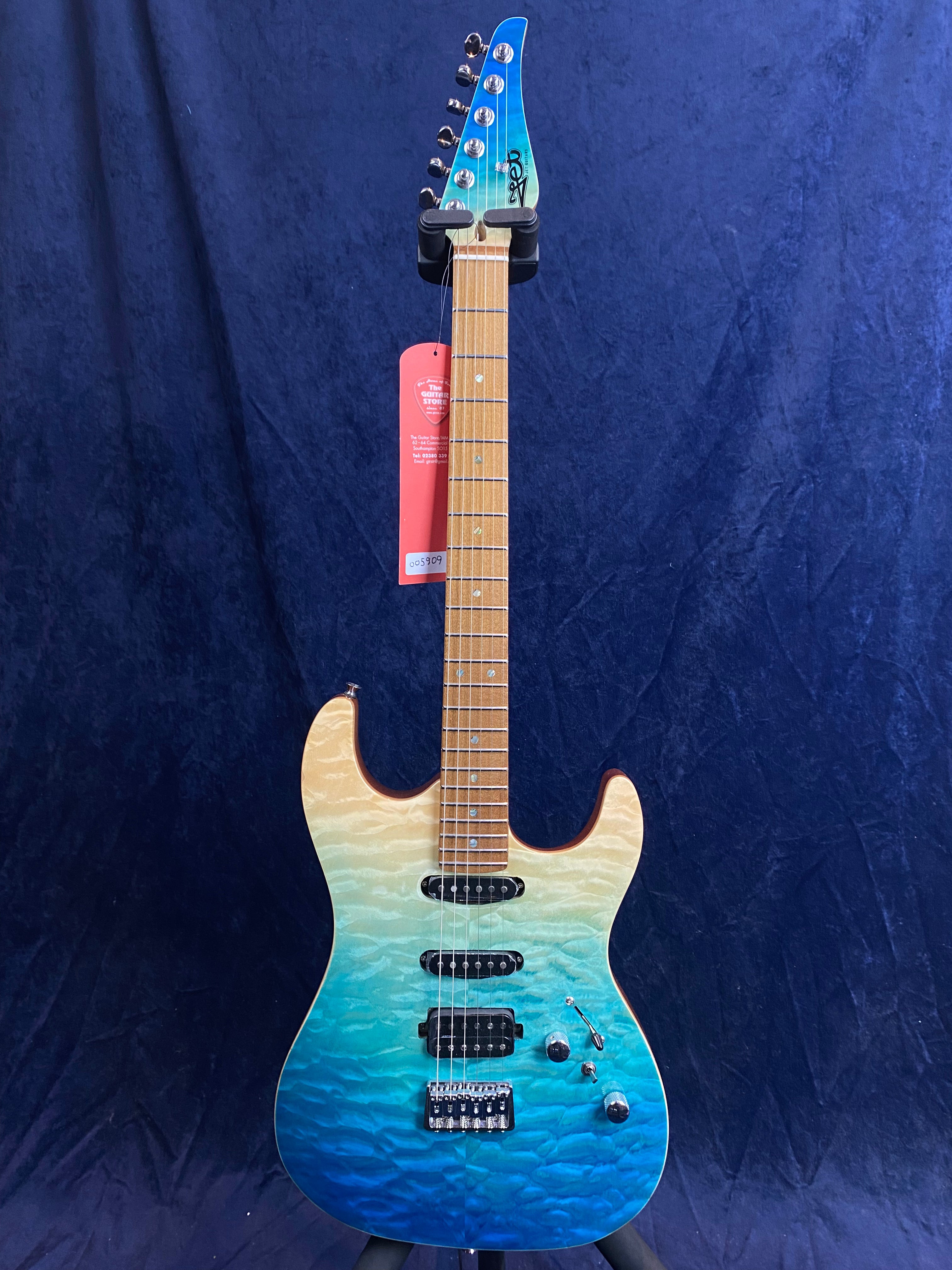 Electric guitar with a quilted top in a blue-to-cream gradient, maple neck, three pickups, and chrome hardware against a dark blue background.