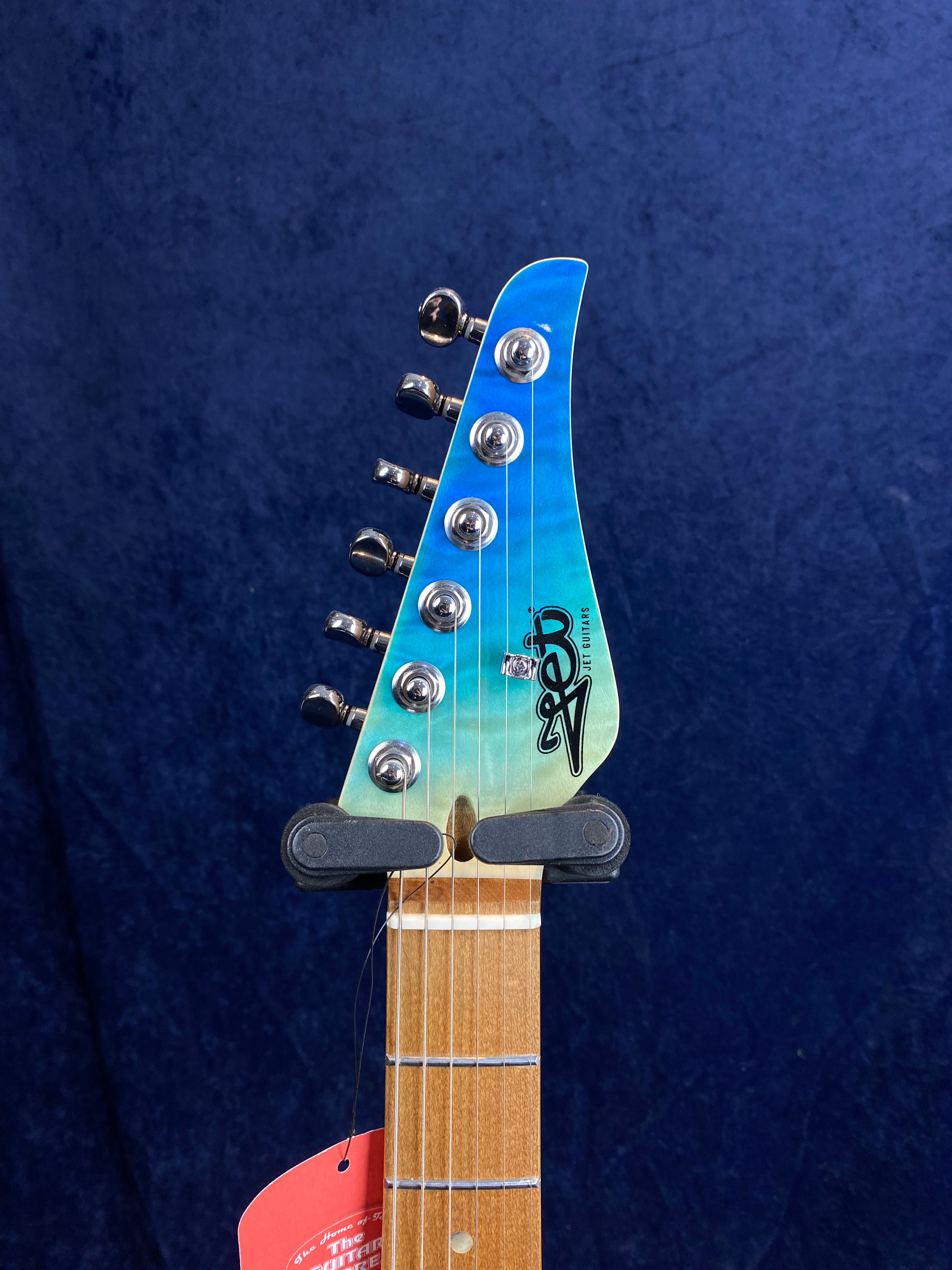 Electric guitar headstock with six tuning pegs, light wood neck, and a blue-green gradient finish with a black logo on the headstock.