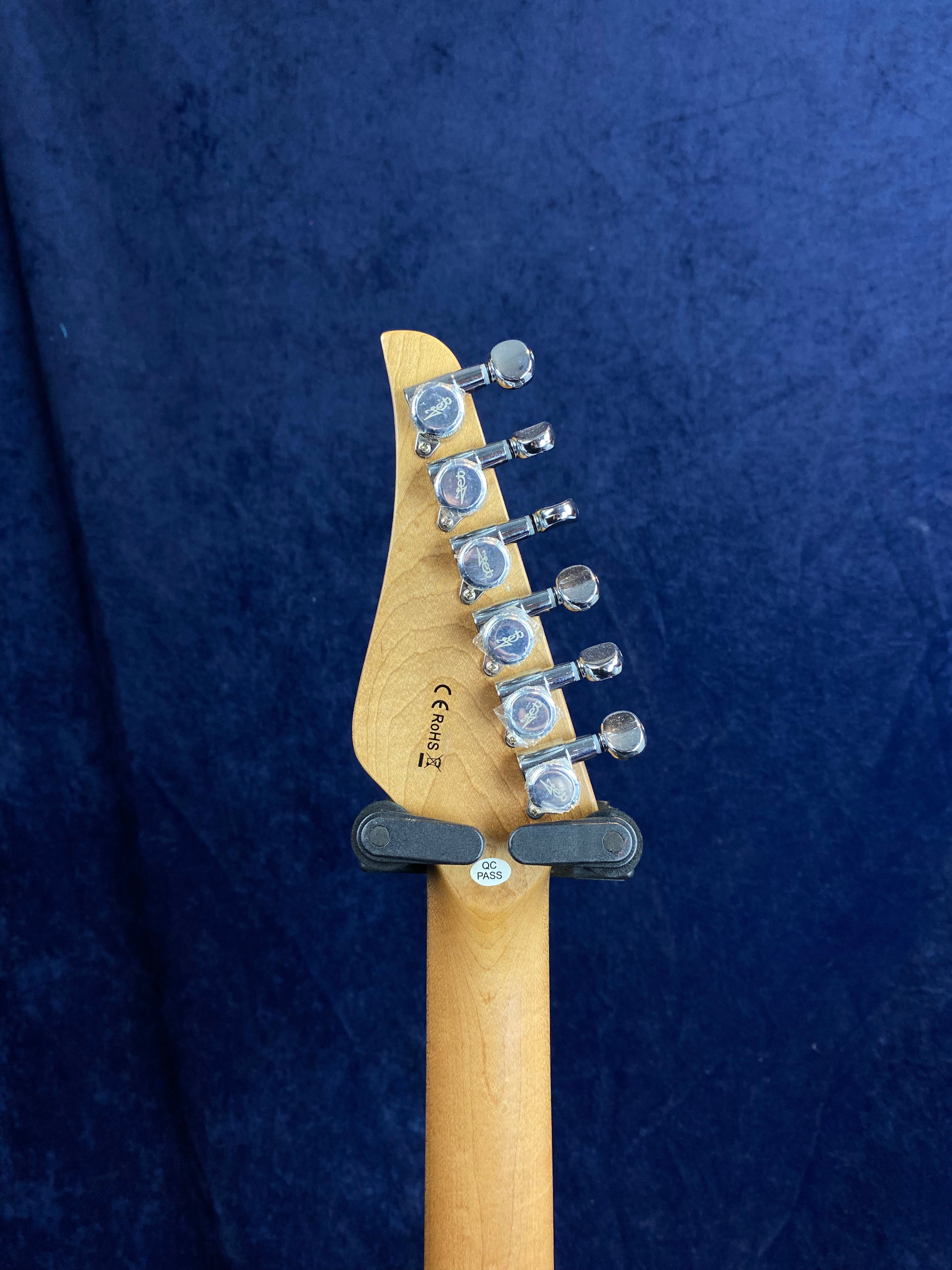 Electric guitar headstock in natural wood with six chrome tuning pegs, viewed from the back against a dark blue background.