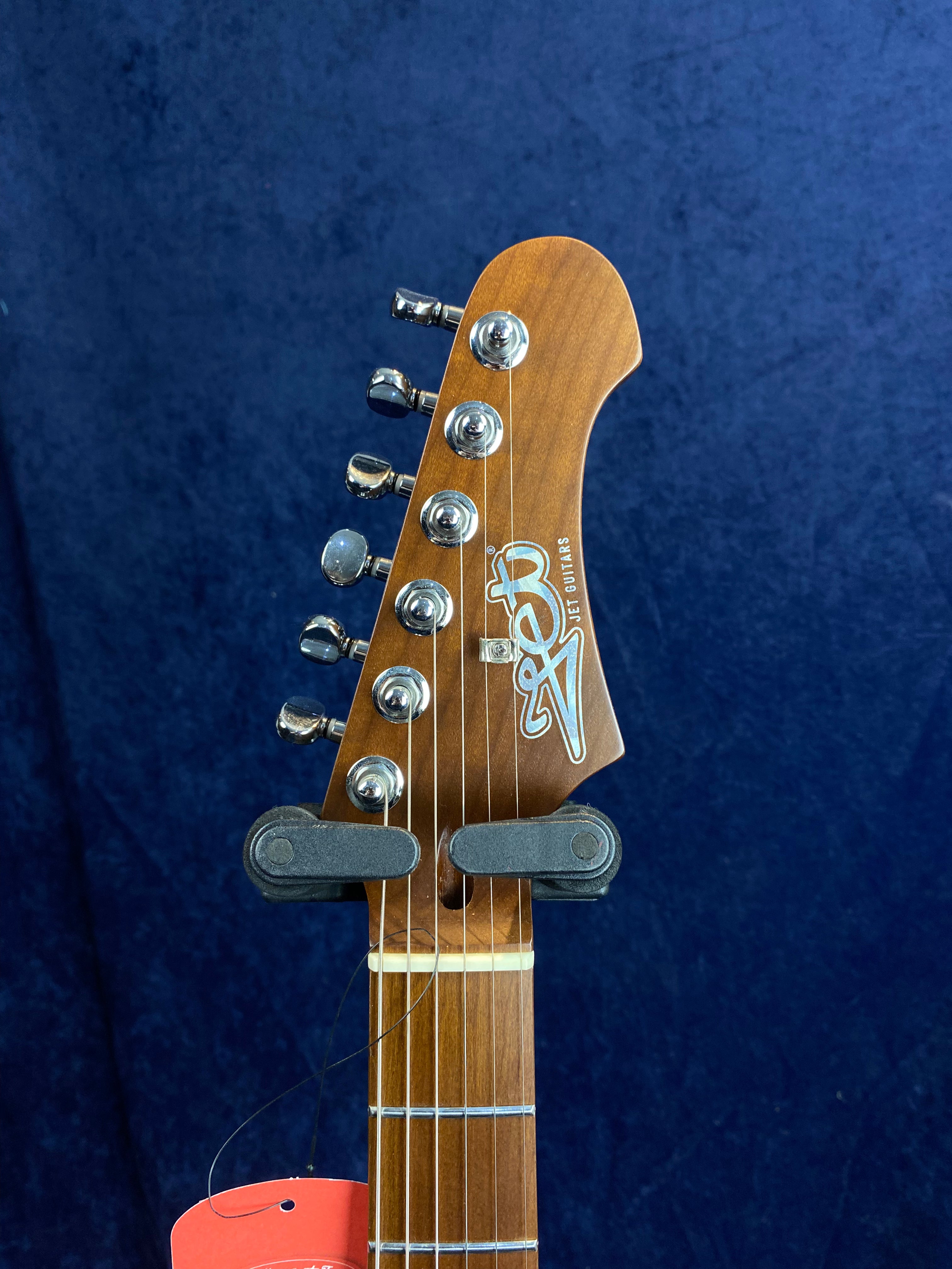 Electric guitar headstock with six metal tuning pegs, brown wood finish, white logo, and part of the fretboard visible against a dark blue background.