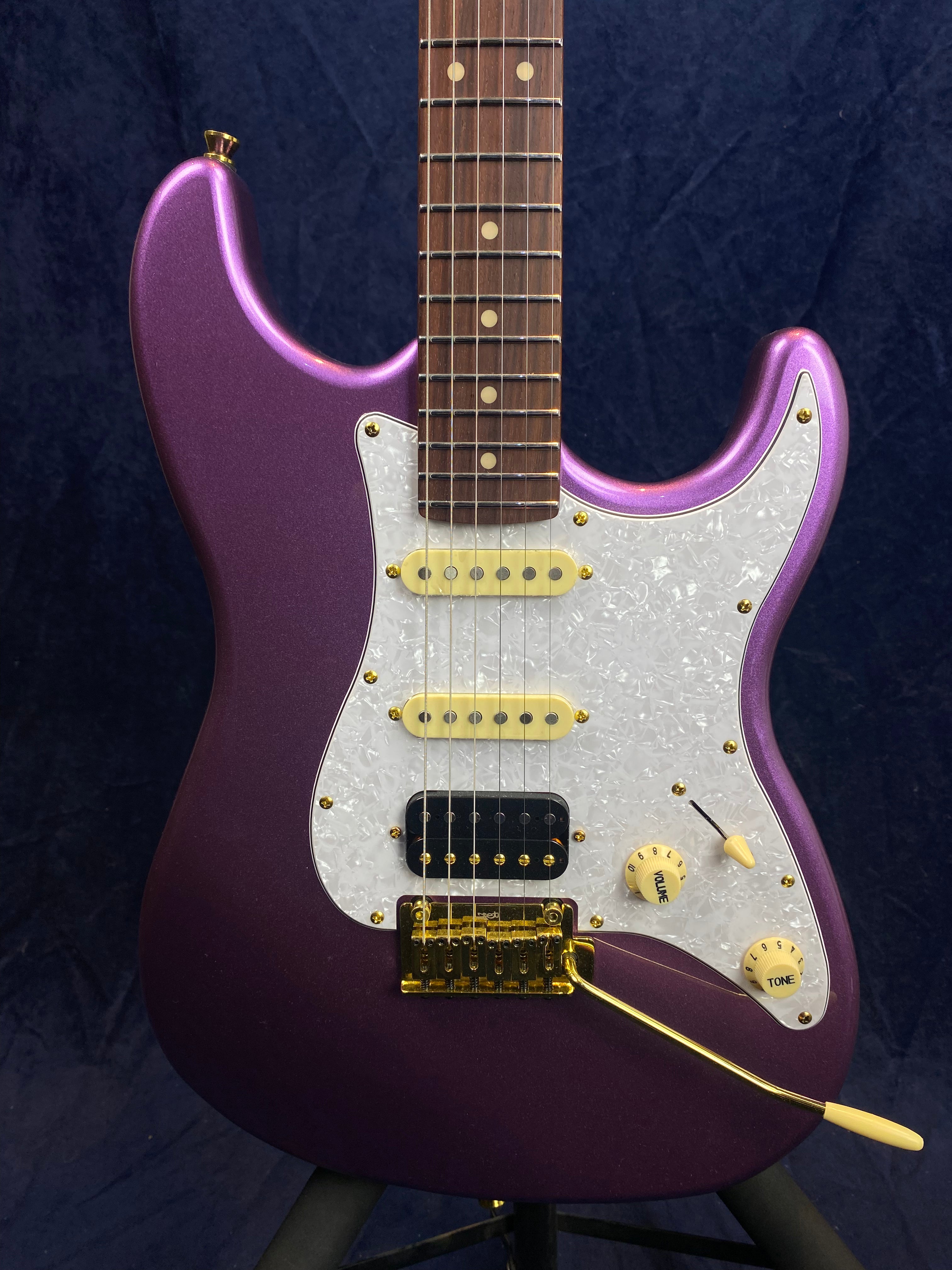 Metallic purple electric guitar with a white pearloid pickguard, gold hardware, three pickups, and cream-colored knobs for volume and tone.