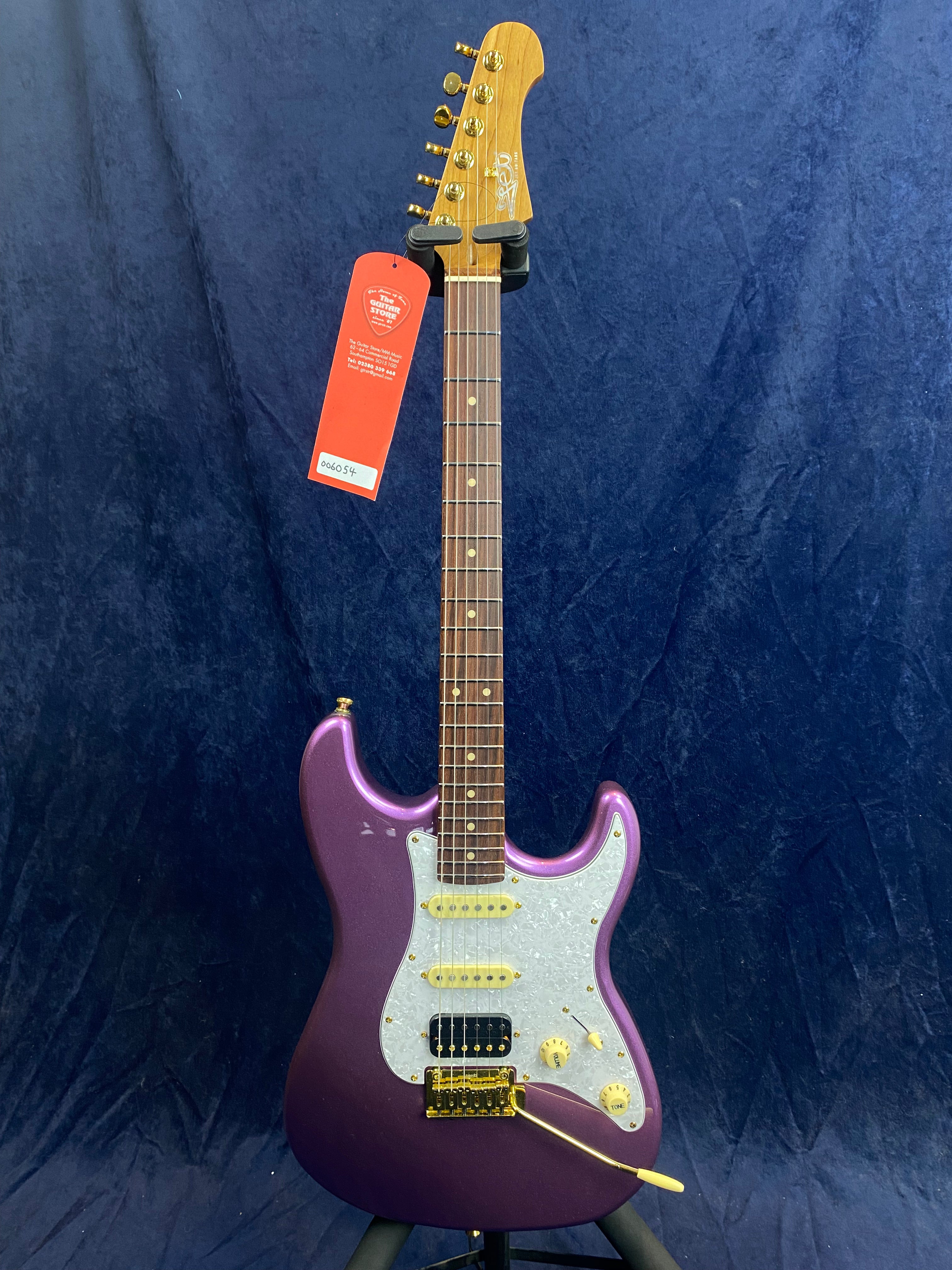 Purple electric guitar with white pickguard, gold hardware, three pickups, maple neck, and a whammy bar, shown against a dark blue background.