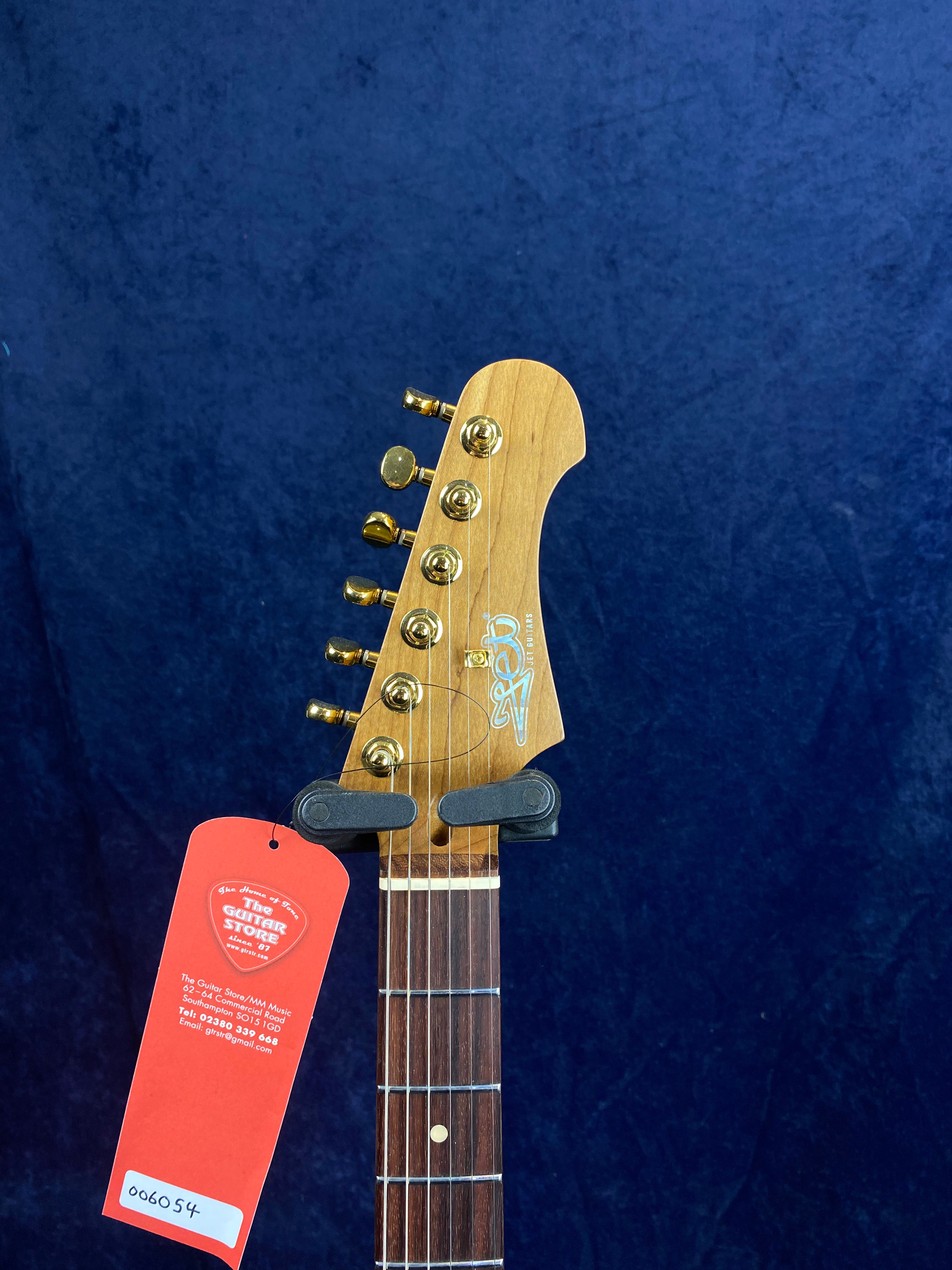 Light brown electric guitar headstock with six gold tuning pegs, logo, and red Guitar Store tag attached below the nut against a dark blue background.