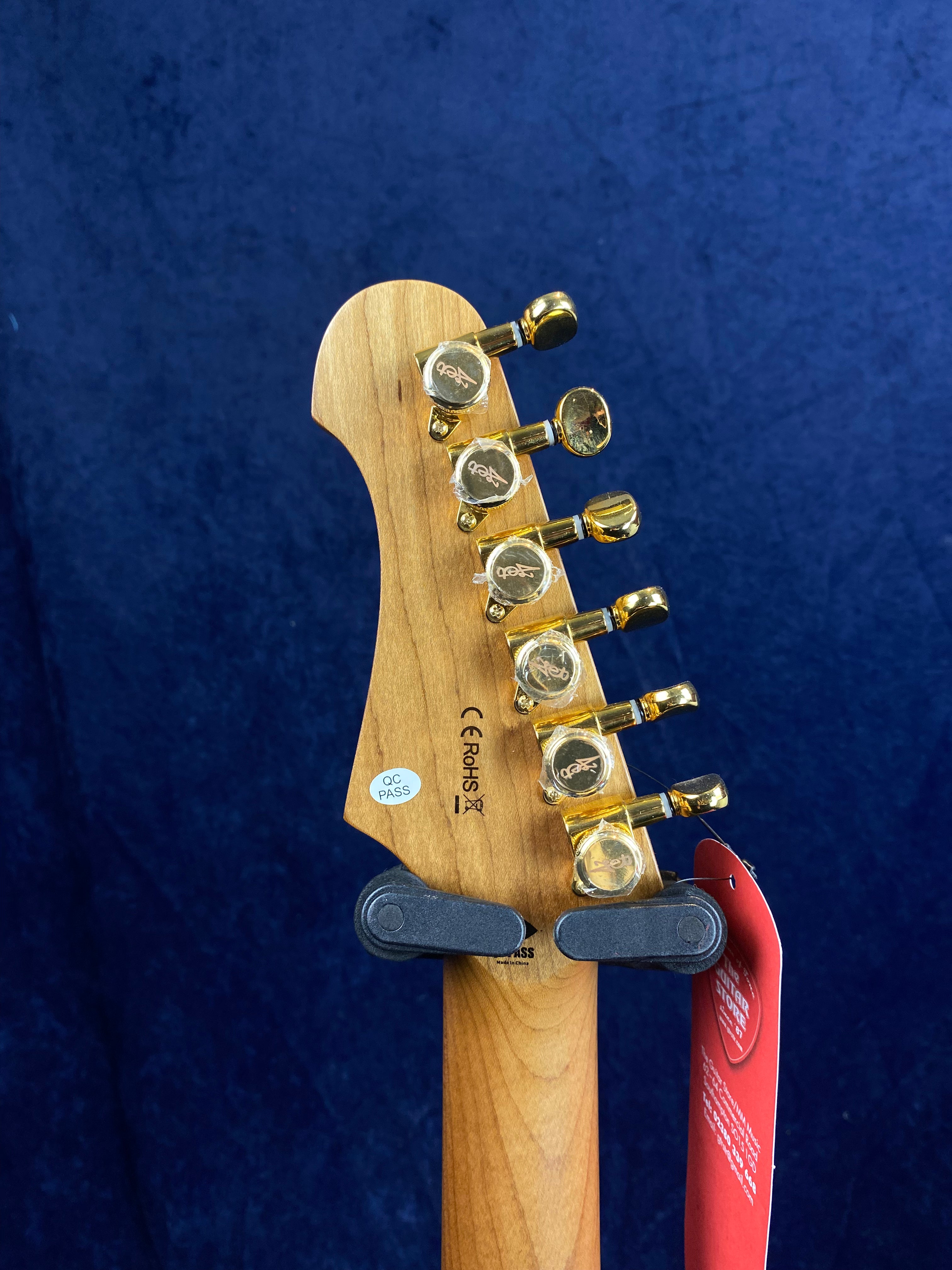 Back of an electric guitar headstock showing six gold tuning pegs, wood grain, certification stickers, and a red hang tag against a dark blue background.