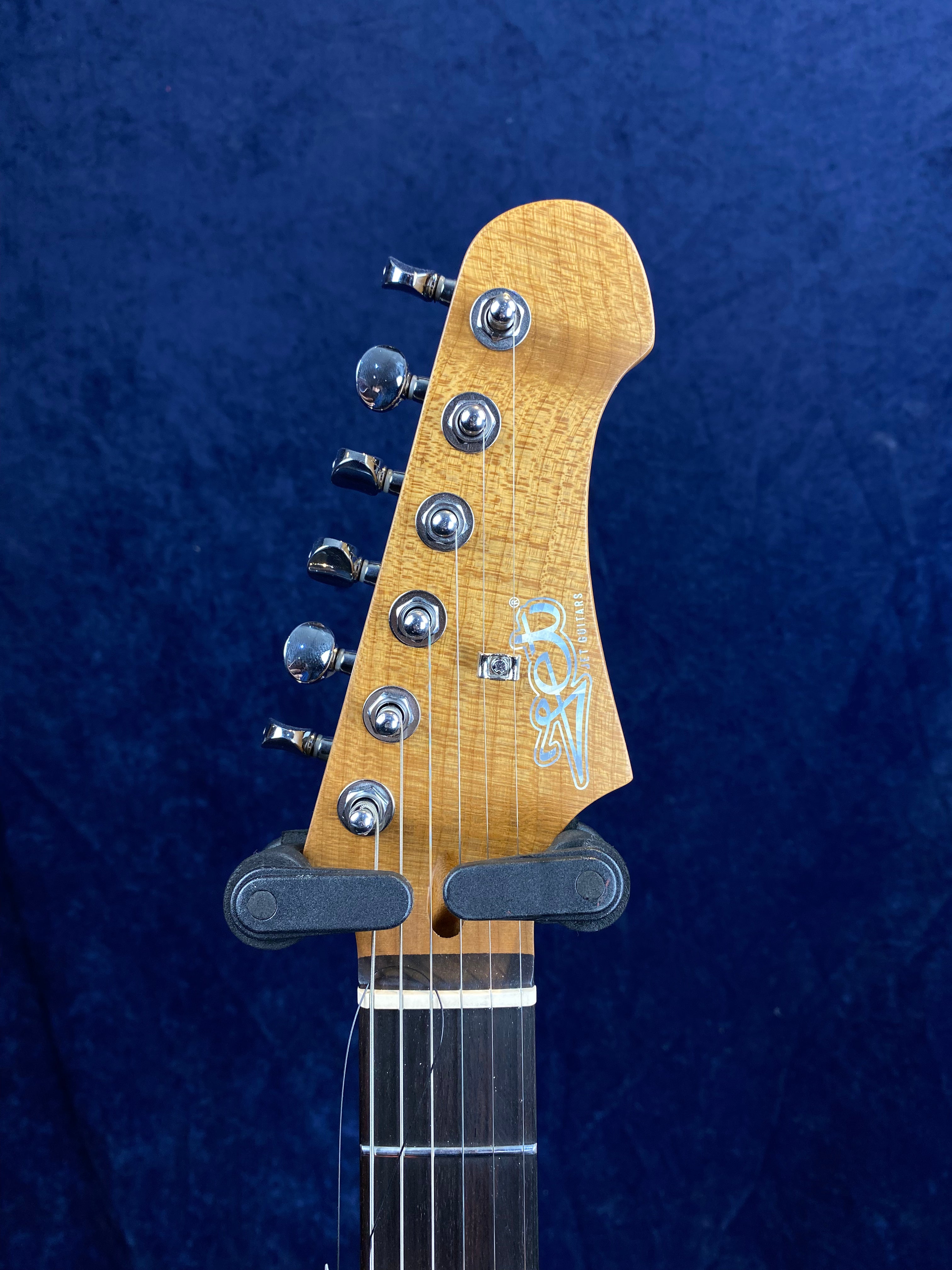 Natural wood finish electric guitar headstock with six chrome tuning pegs and a white logo, against a dark blue background.