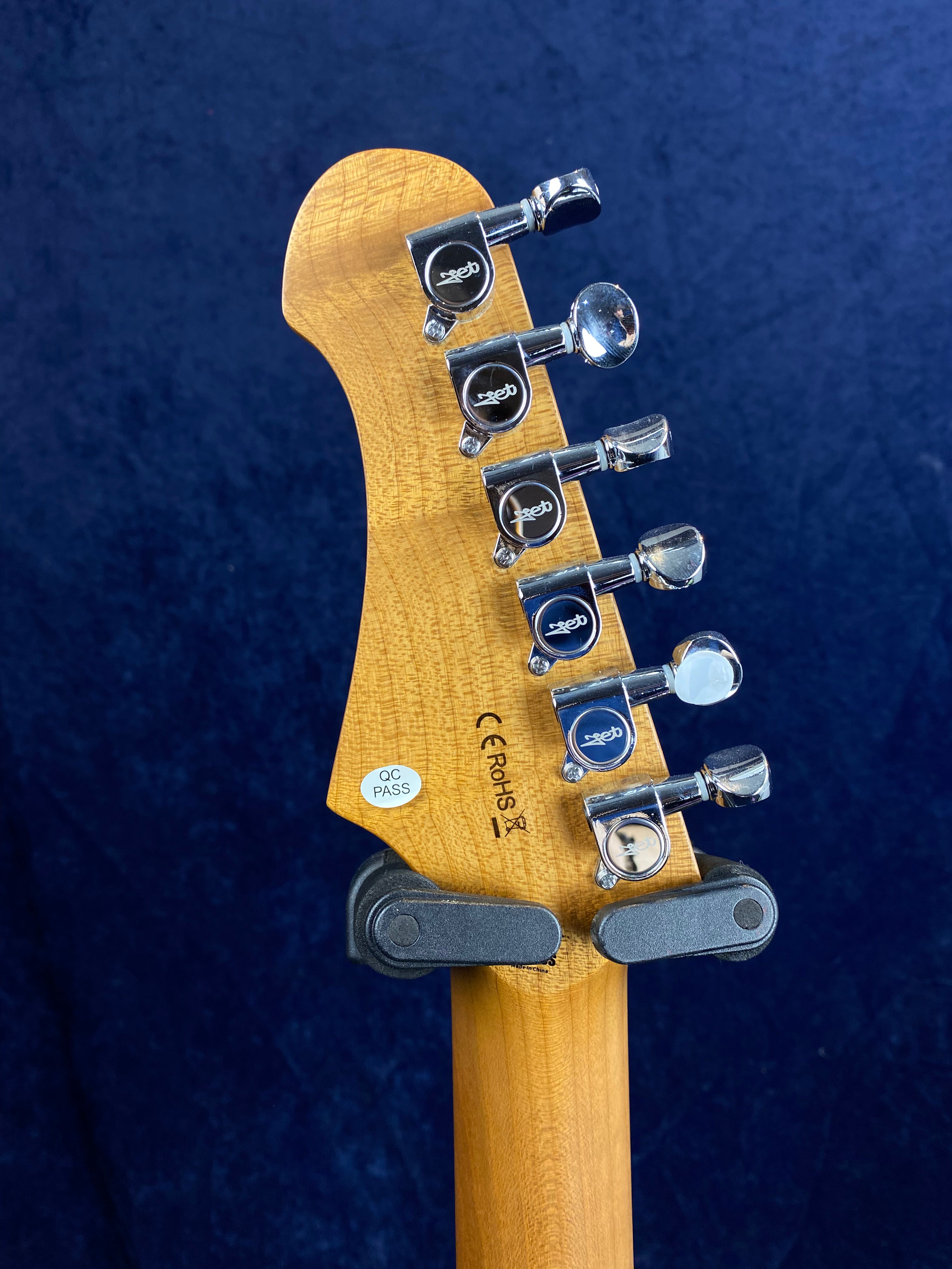 Six chrome tuning pegs on a light wood electric guitar headstock, with certification stickers and a wall mount cradle holding the neck.