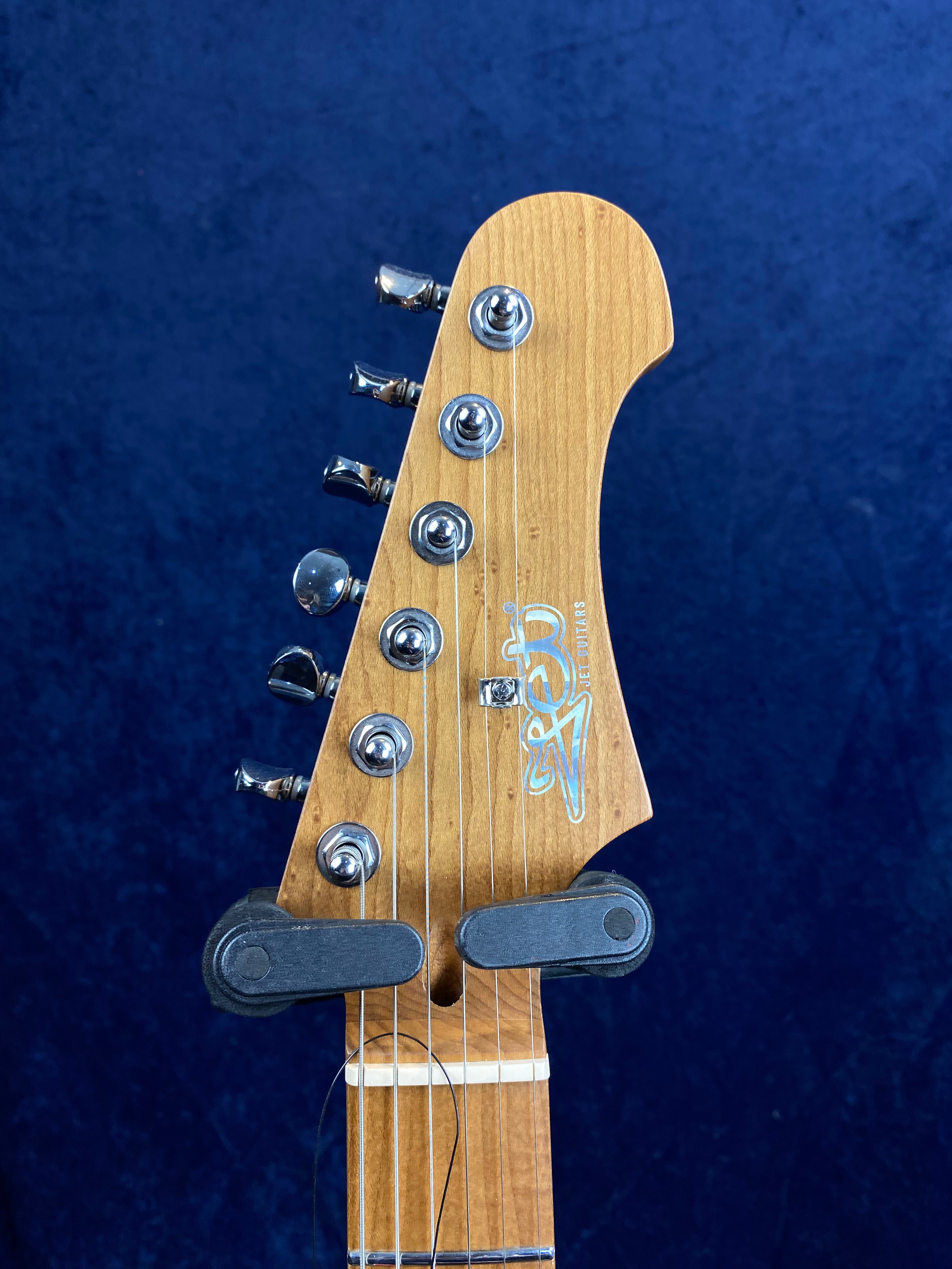 Natural wood electric guitar headstock with six metal tuning pegs, logo, and a capodastro clipped just above the nut, against a blue background.