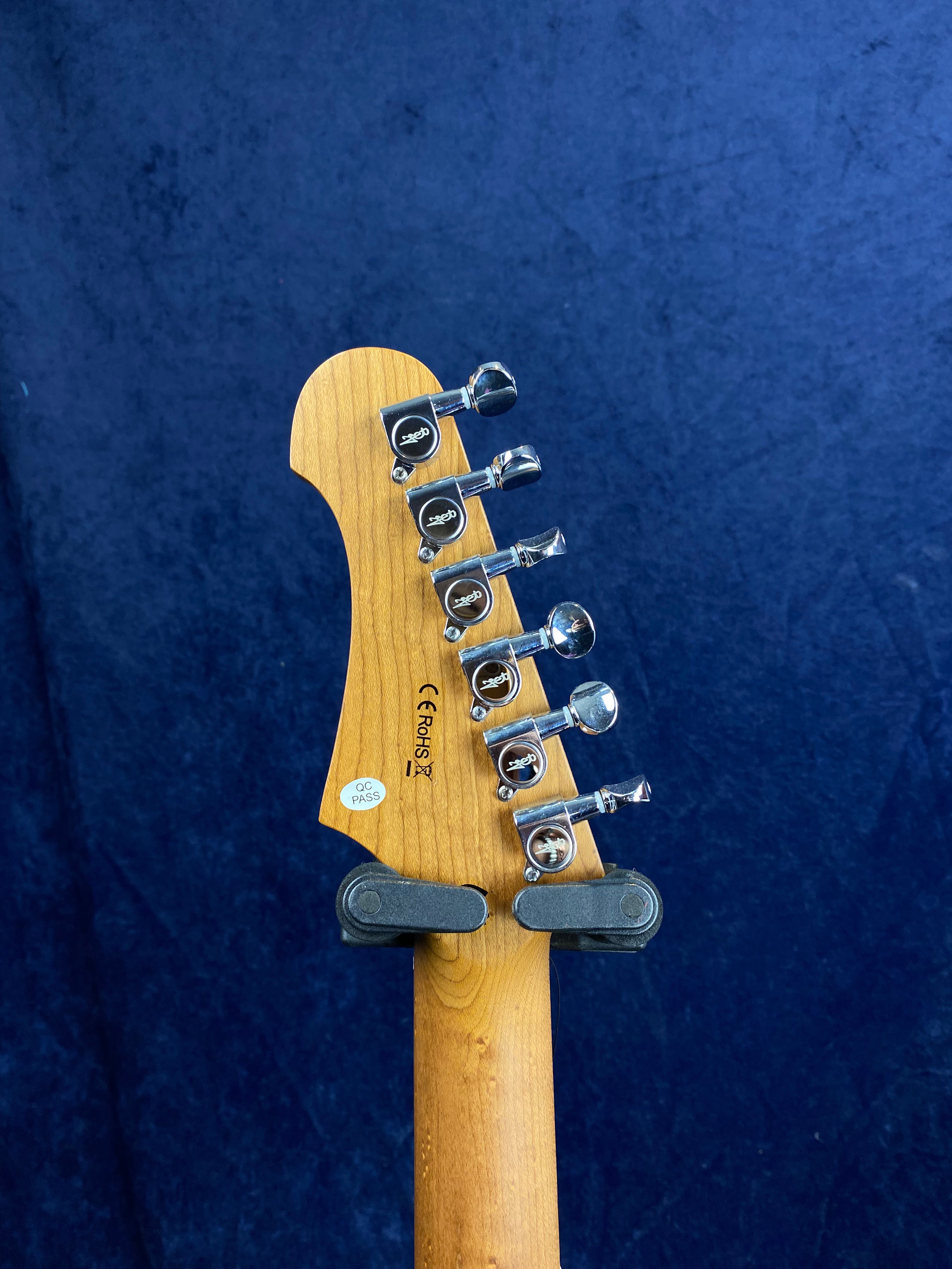 Electric guitar headstock showing six chrome tuning pegs, certification markings, and a QC pass sticker against a dark blue background.