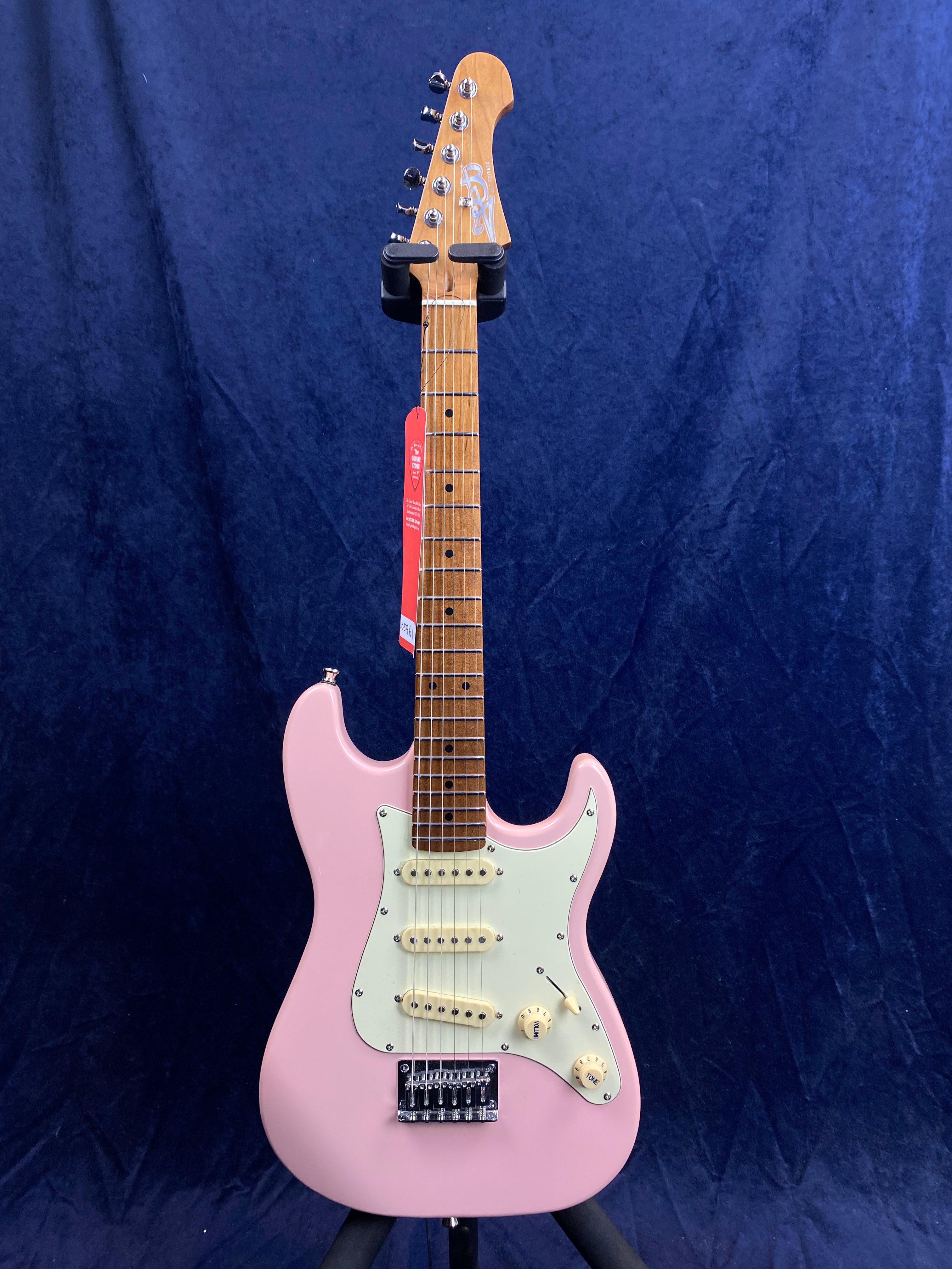 Pink electric guitar with three white pickups, cream knobs, a white pickguard, and a maple neck, displayed upright on a stand against a dark blue background.