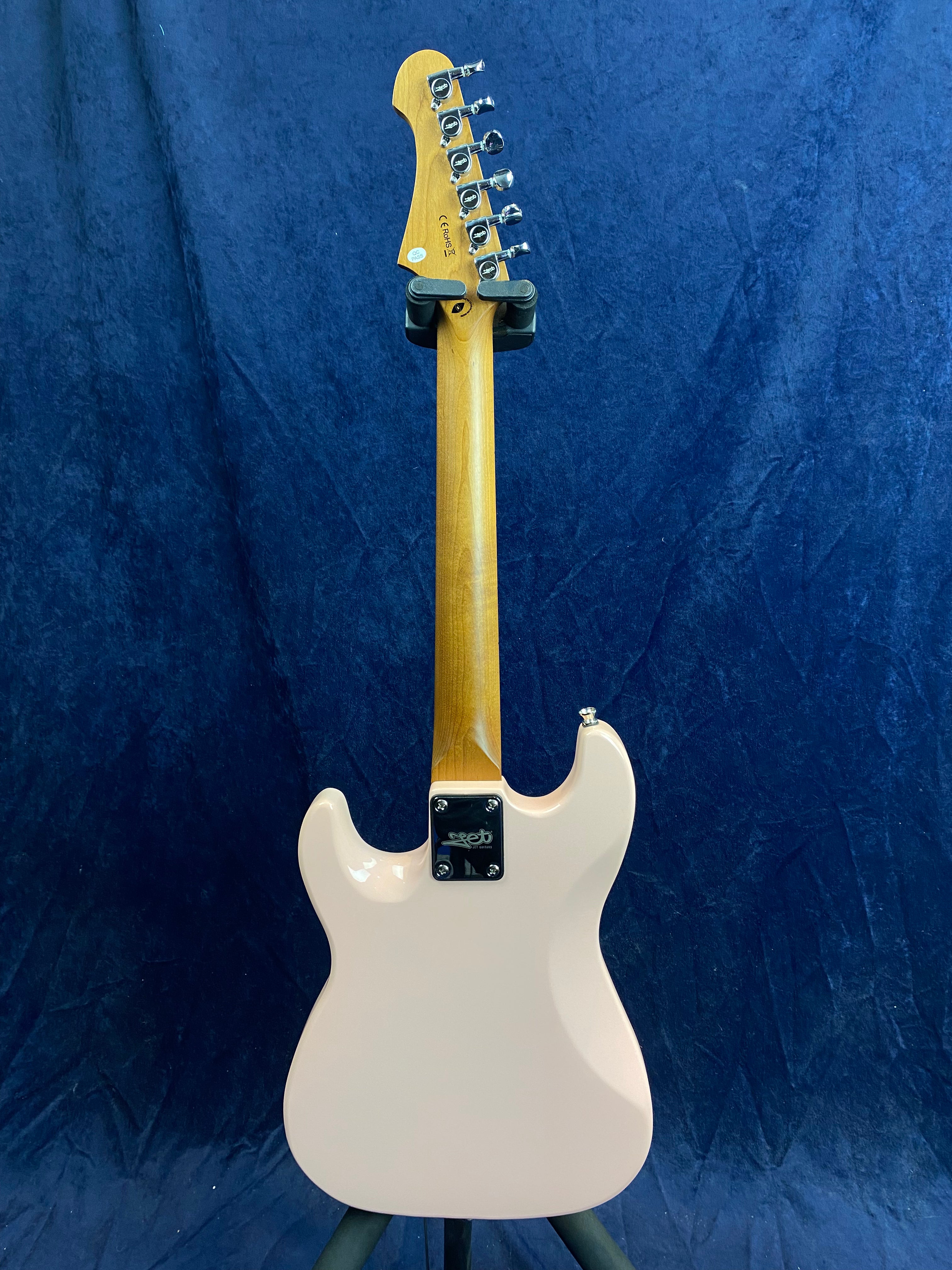 Electric guitar shown from the back, with a light pink body, natural wood neck, and six tuning pegs on the headstock, standing against a blue background.