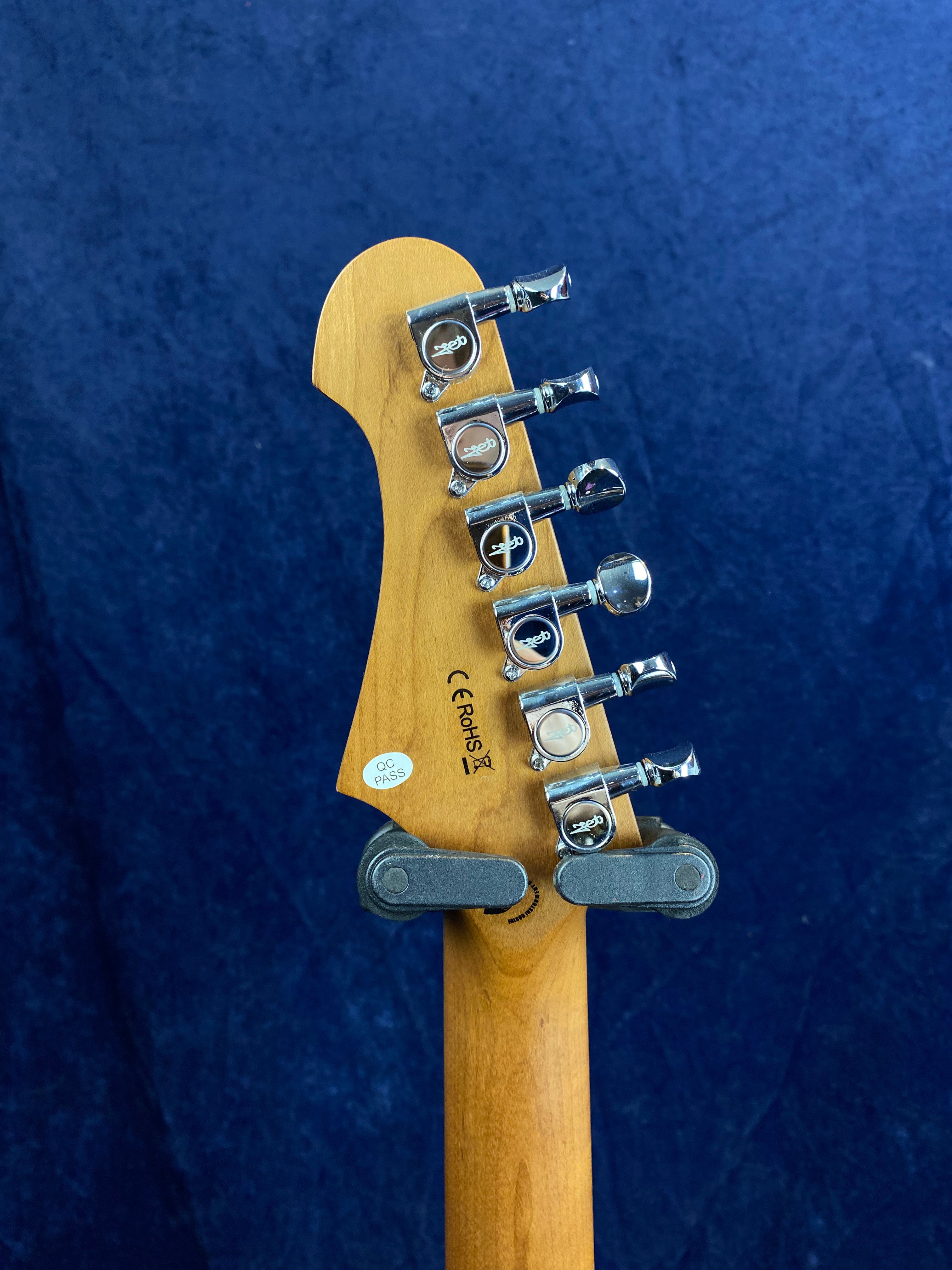 Back of an electric guitar headstock showing six chrome tuning pegs, certification stickers, and part of a wall hanger against a blue background.