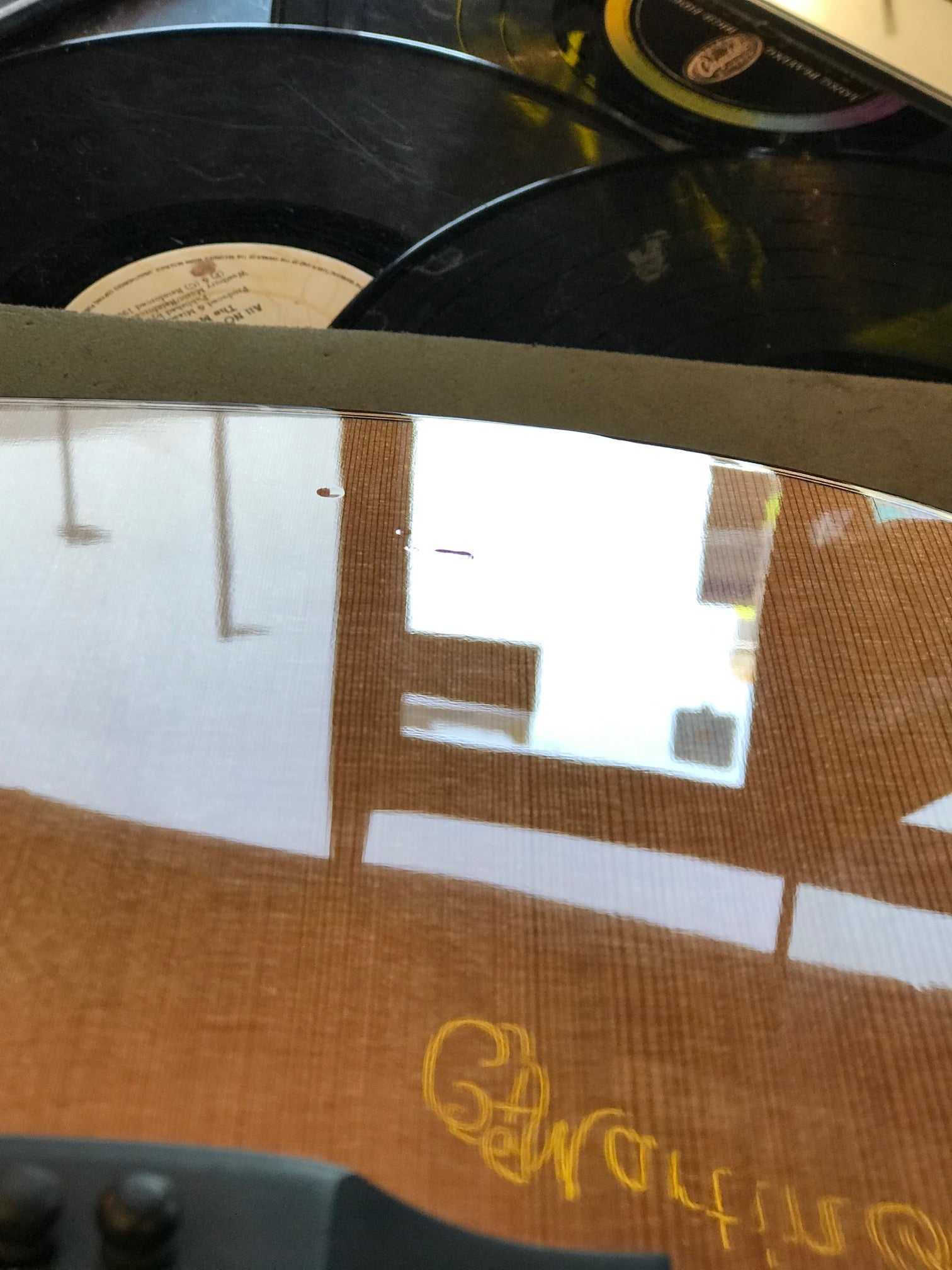A stack of vinyl records is seen reflected on the glossy wooden surface of an acoustic guitar with some yellow writing visible on the guitar's body.