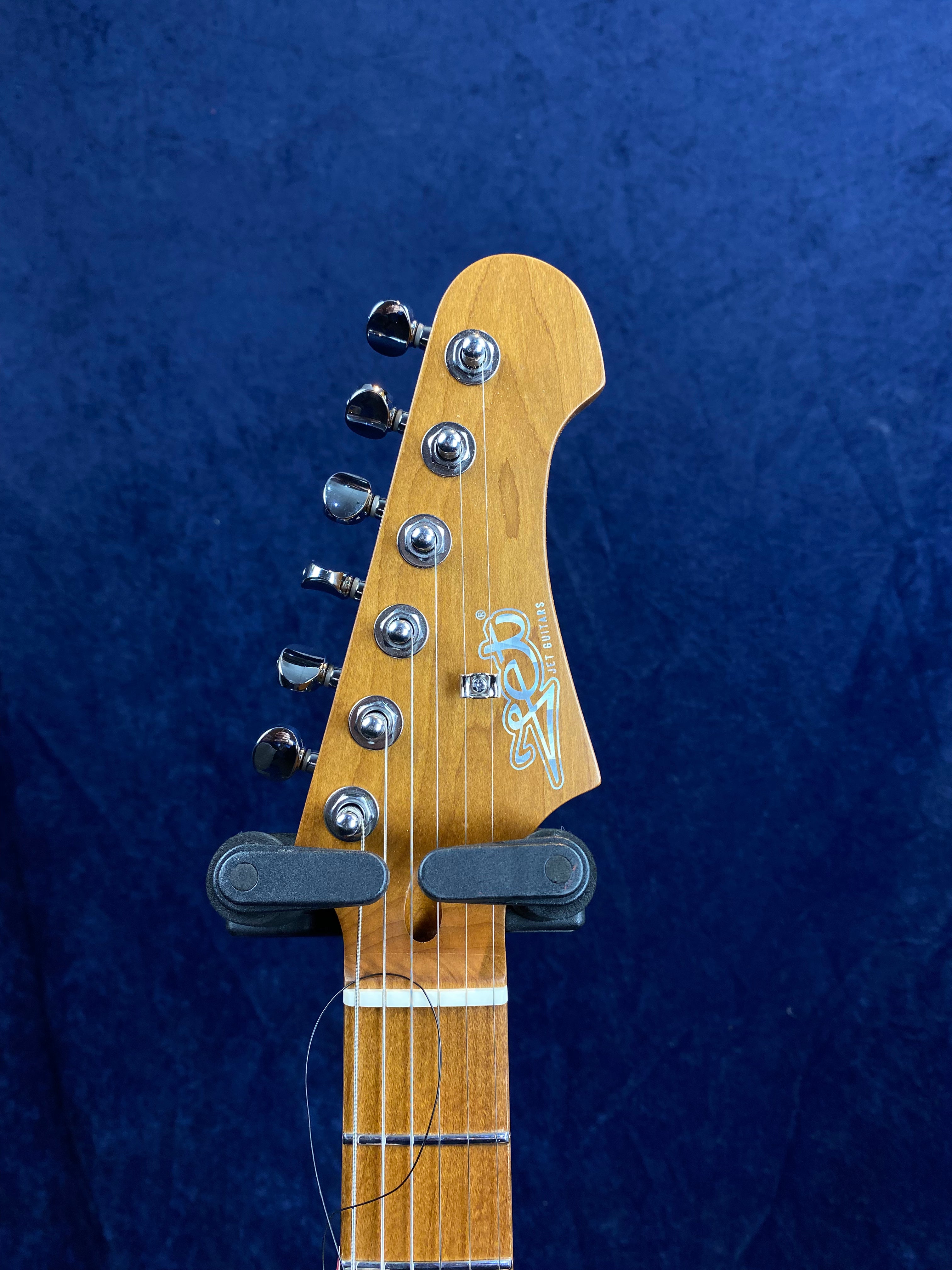 Six-string Jet Guitars headstock with chrome tuning pegs, wood grain finish, and white Jet logo, set against a dark blue background.