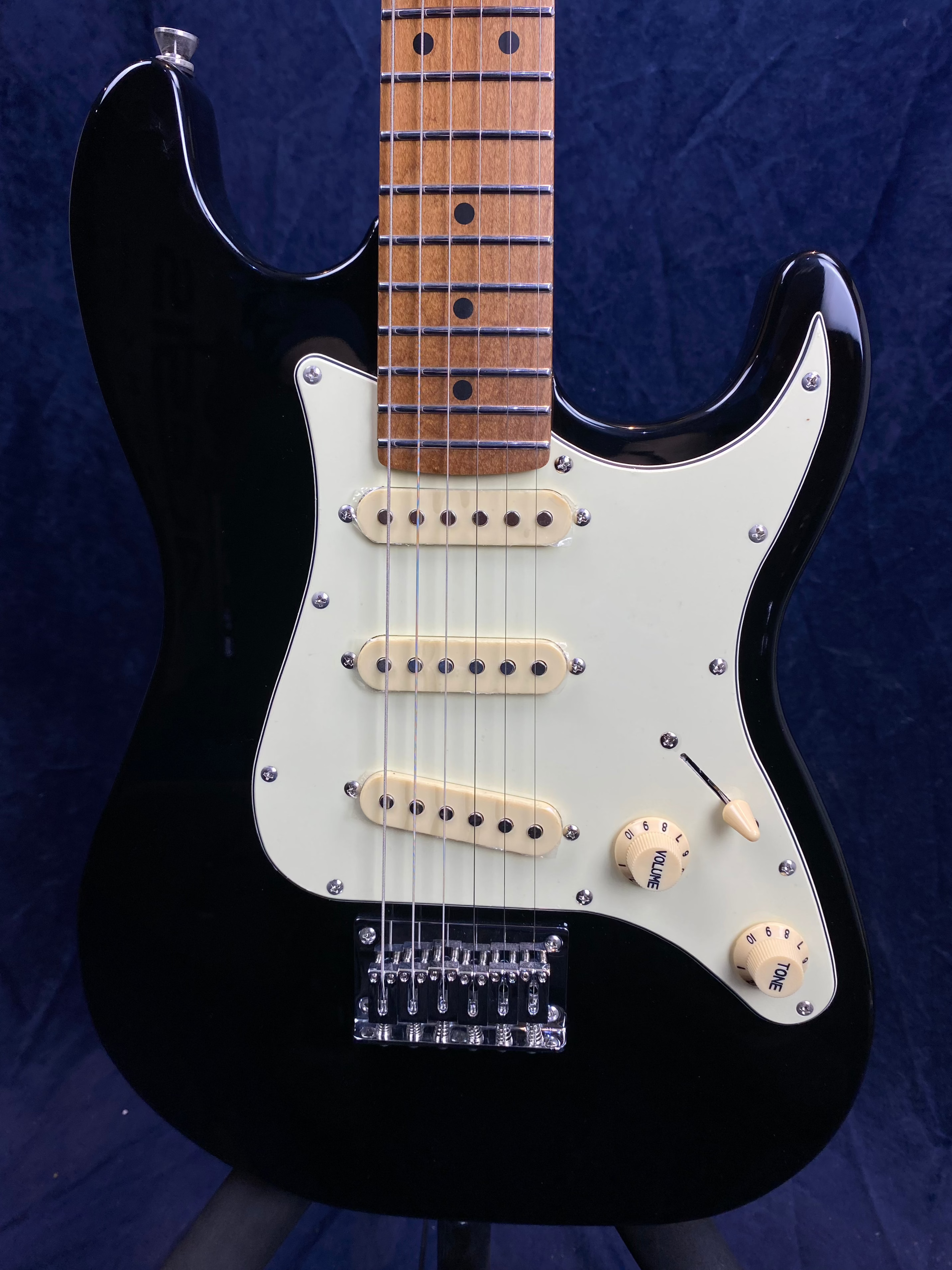 Black electric guitar with white pickguard, three cream single-coil pickups, two cream knobs labeled volume and tone, and a lever switch.