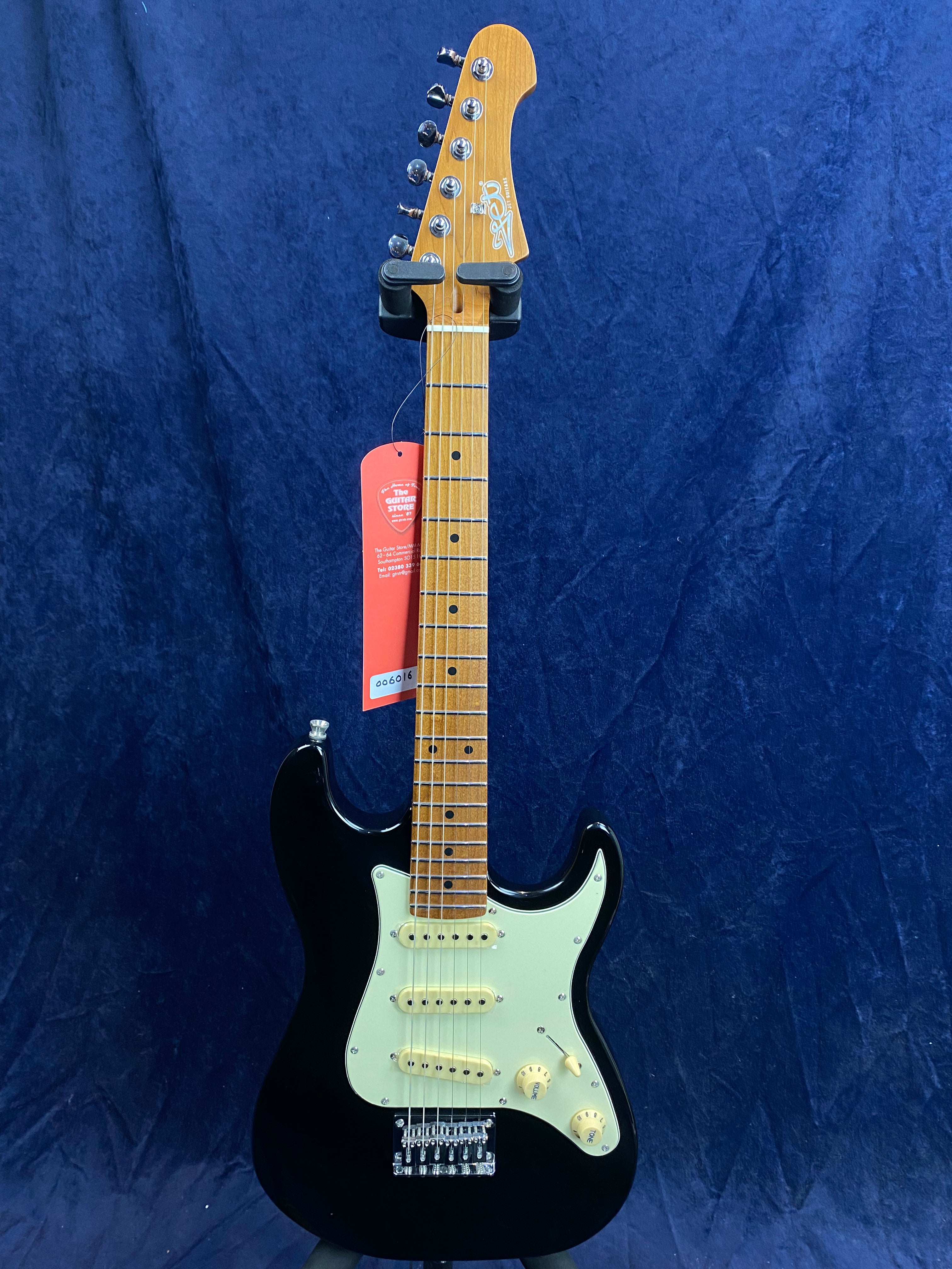 Black electric guitar with a light wood neck, white pickguard, three single-coil pickups, and control knobs, hanging on a display against a blue background.