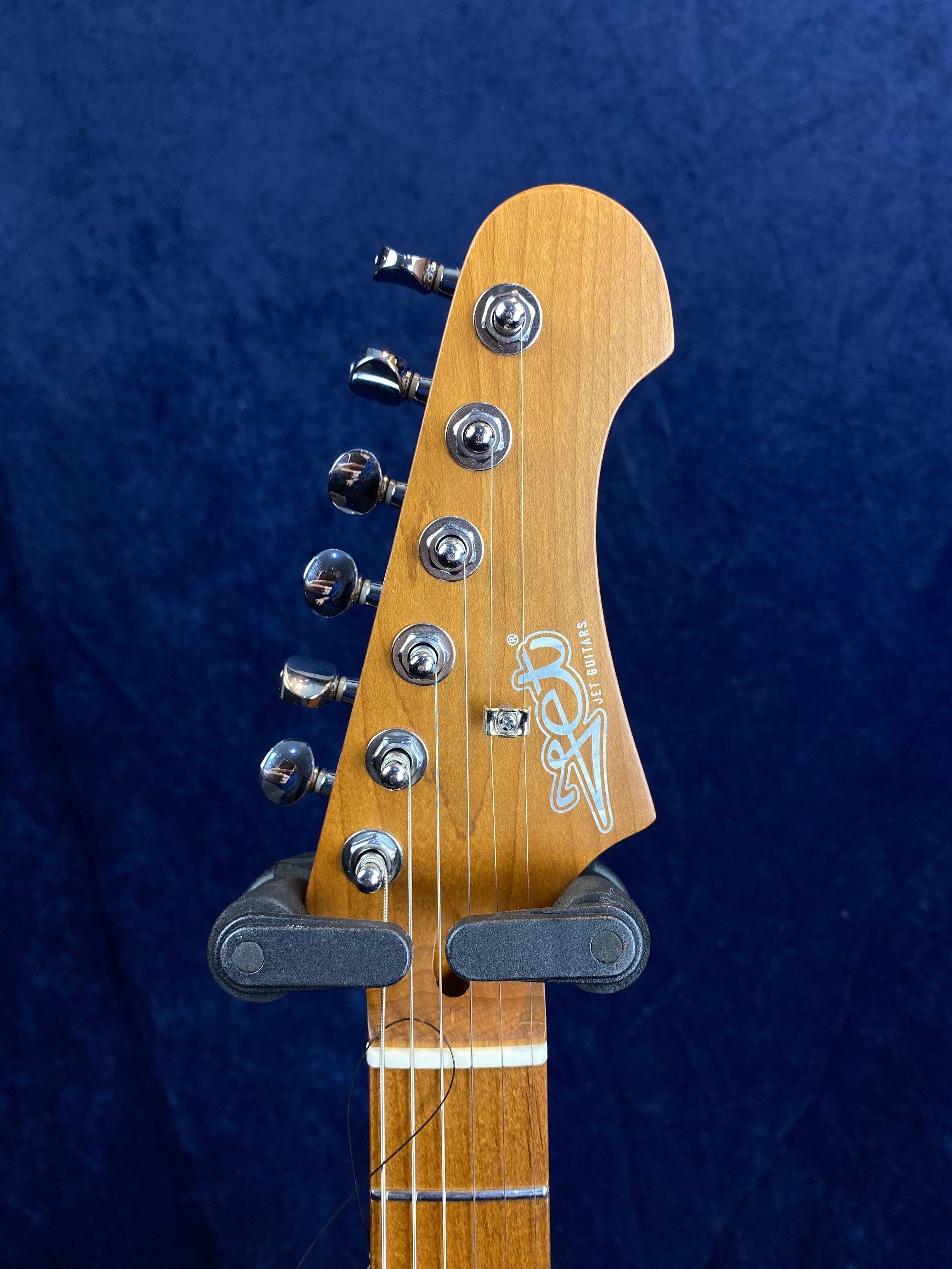 Six-string electric guitar headstock with chrome tuning pegs, wood finish, and “Jet Guitars” logo, against a dark blue background.