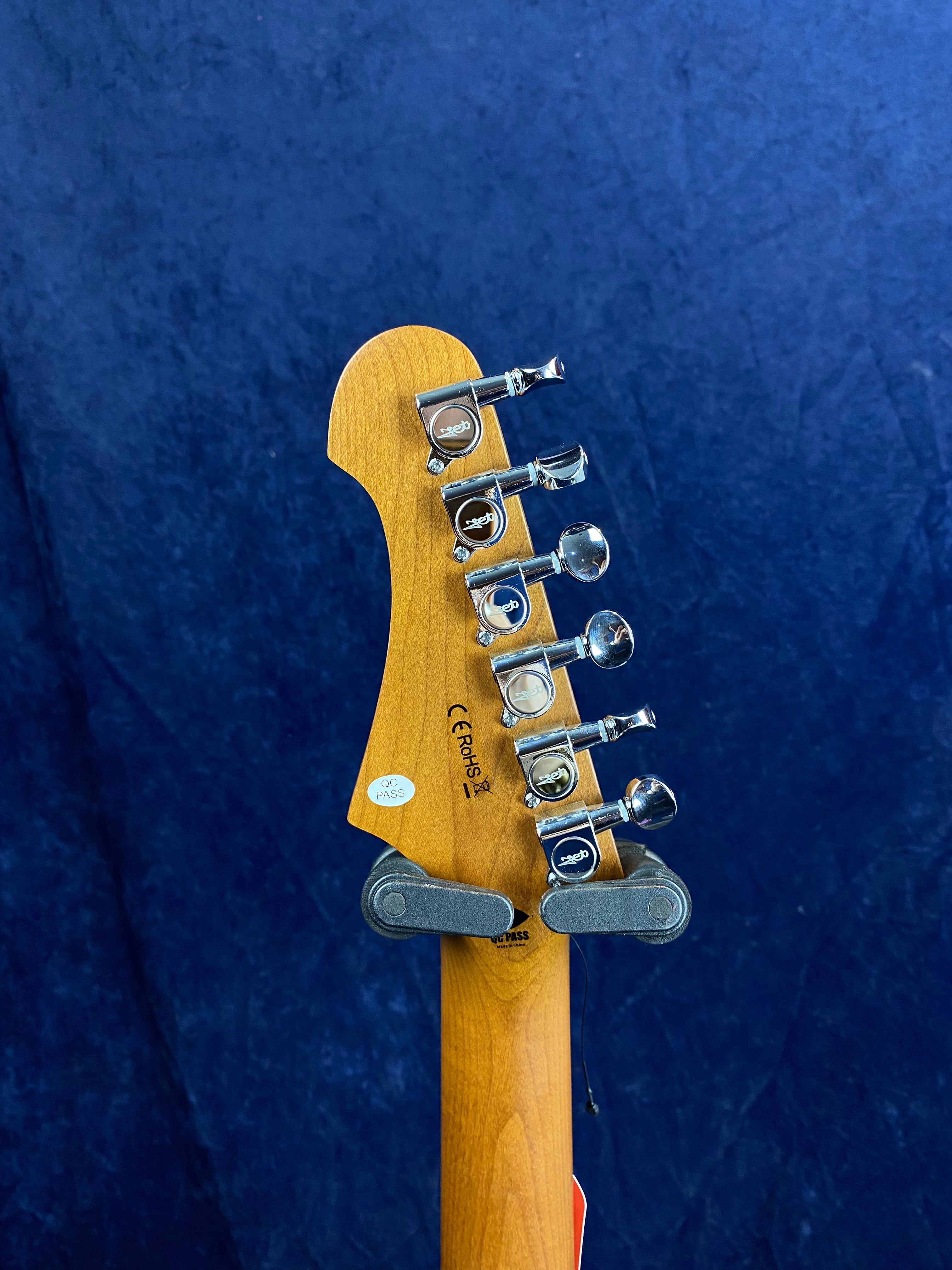 Back of an electric guitar headstock showing six chrome tuning pegs, product markings, and a QC PASS sticker against a dark blue background.