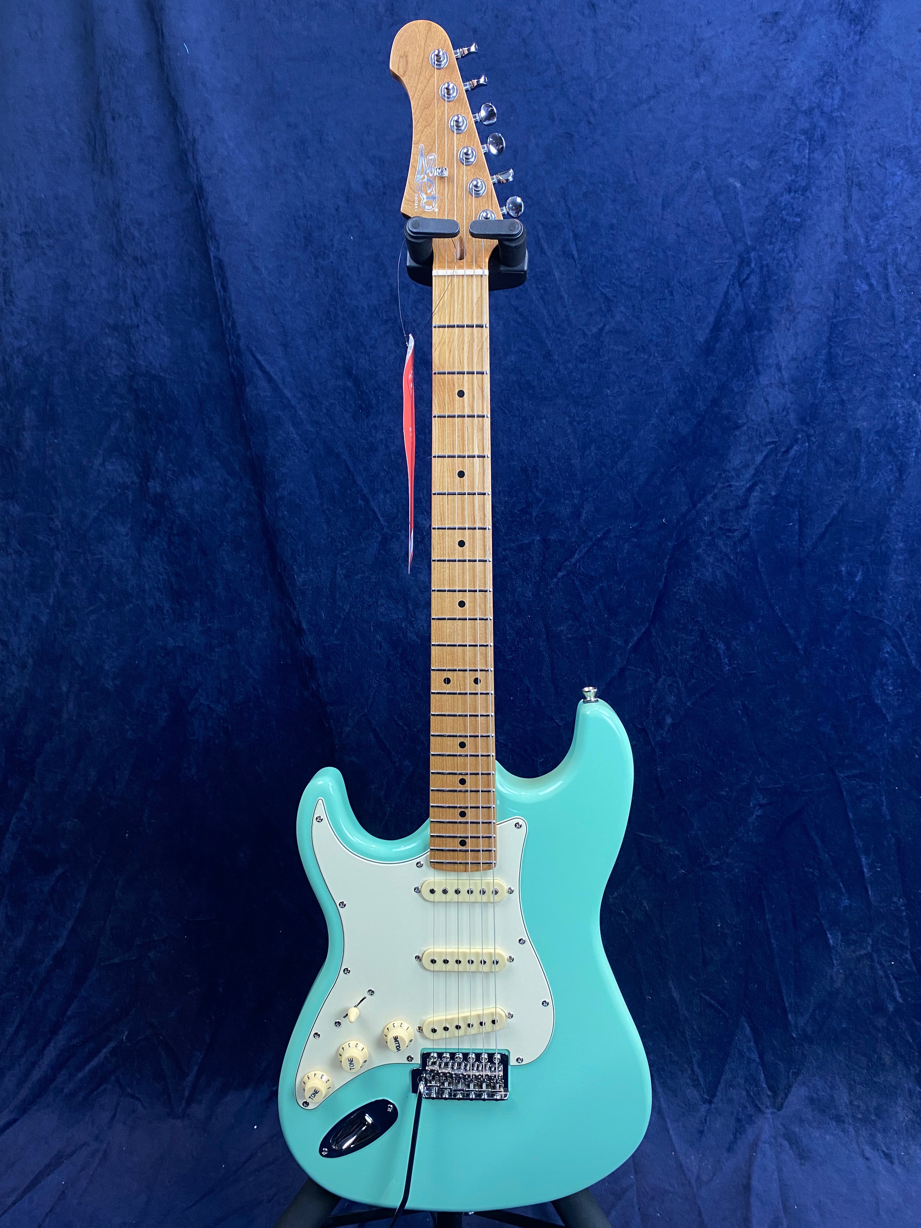 Aqua blue electric guitar with a white pickguard, three single-coil pickups, maple neck, and six tuning pegs against a dark blue background.