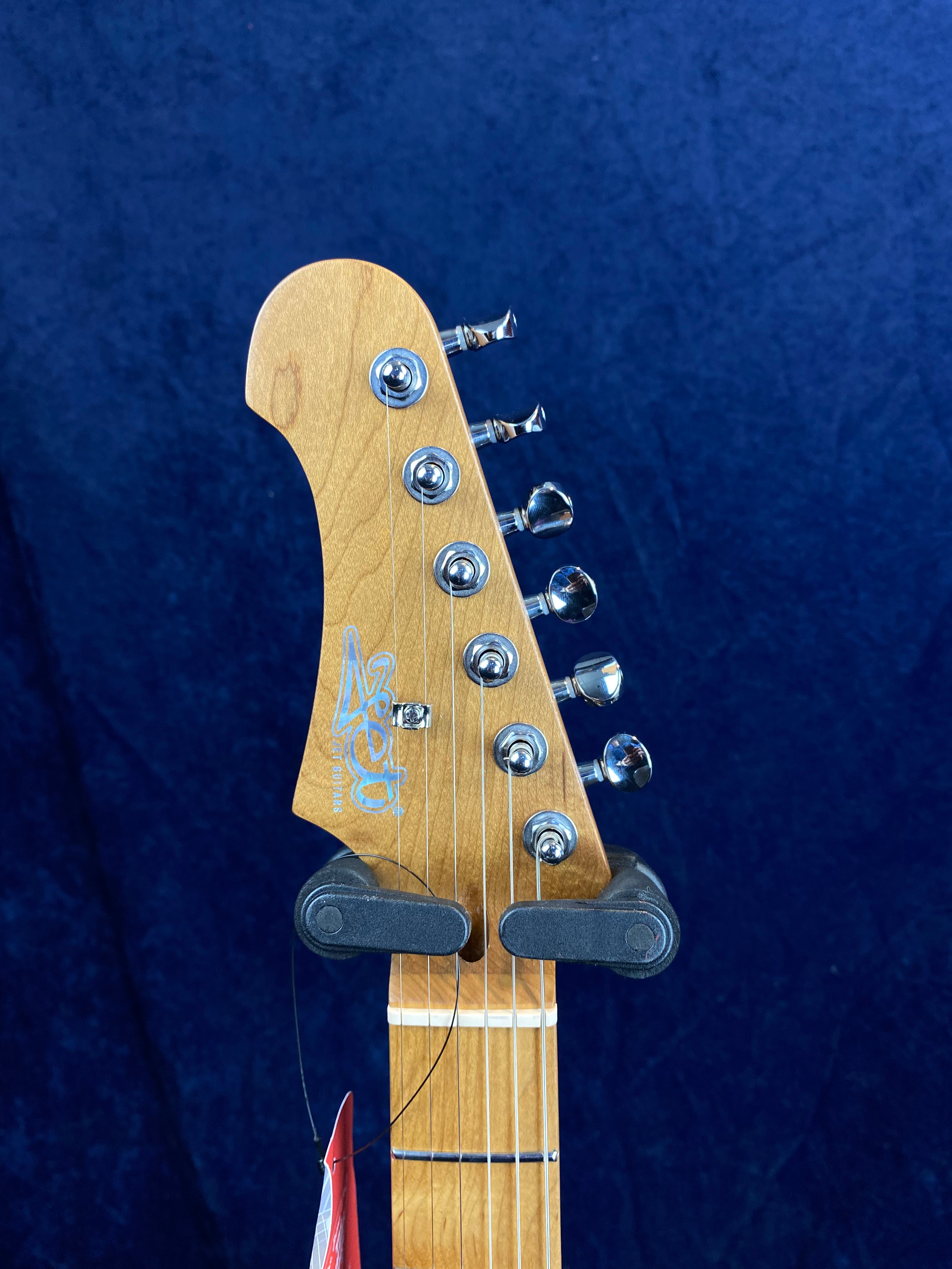Light brown electric guitar headstock with six chrome tuning pegs, string guide, and a visible logo reading "Asher Guitars" on a dark blue background.