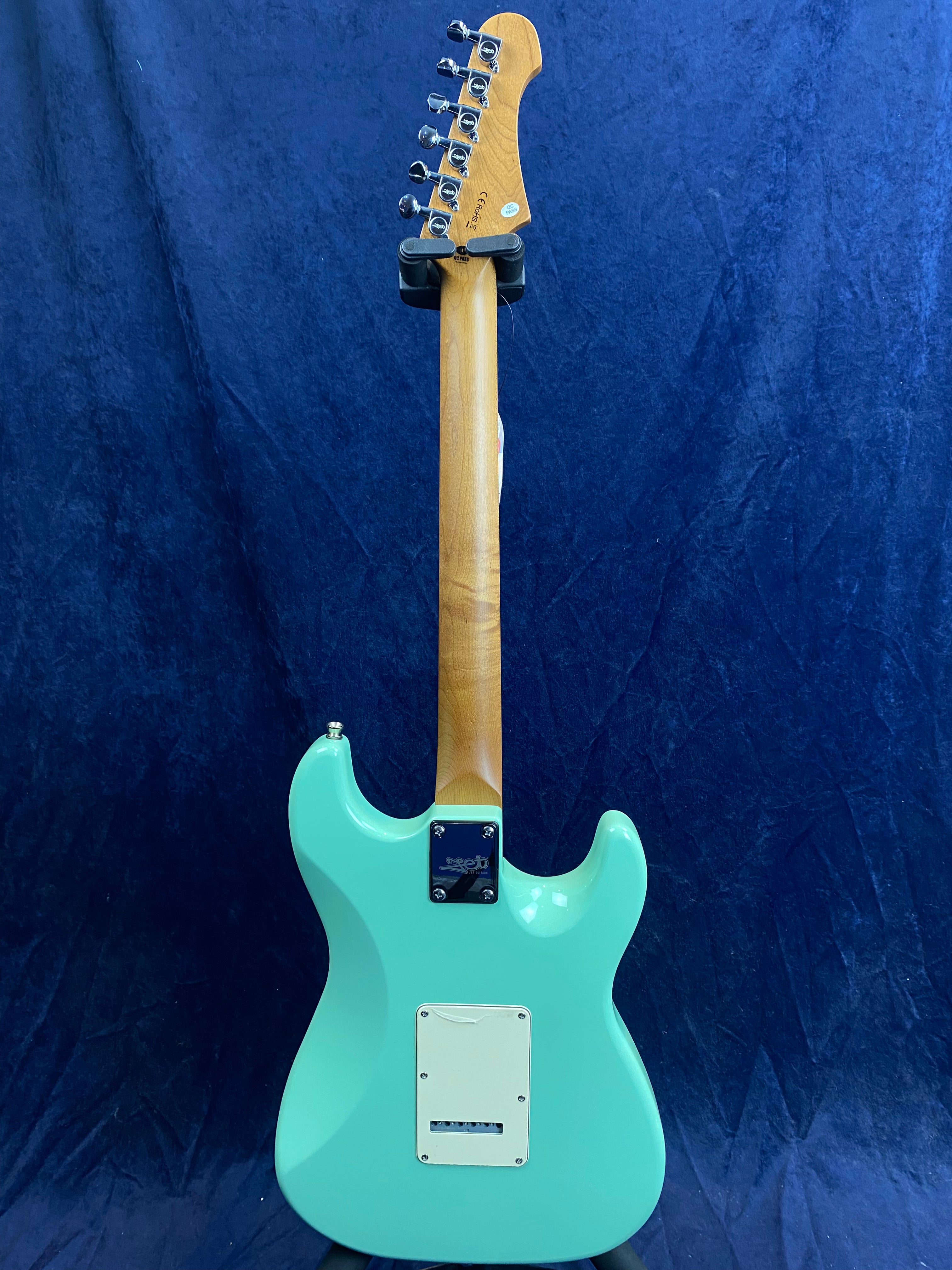 Electric guitar with a light turquoise body, natural wood neck, and chrome tuning pegs, shown from the back against a dark blue background.