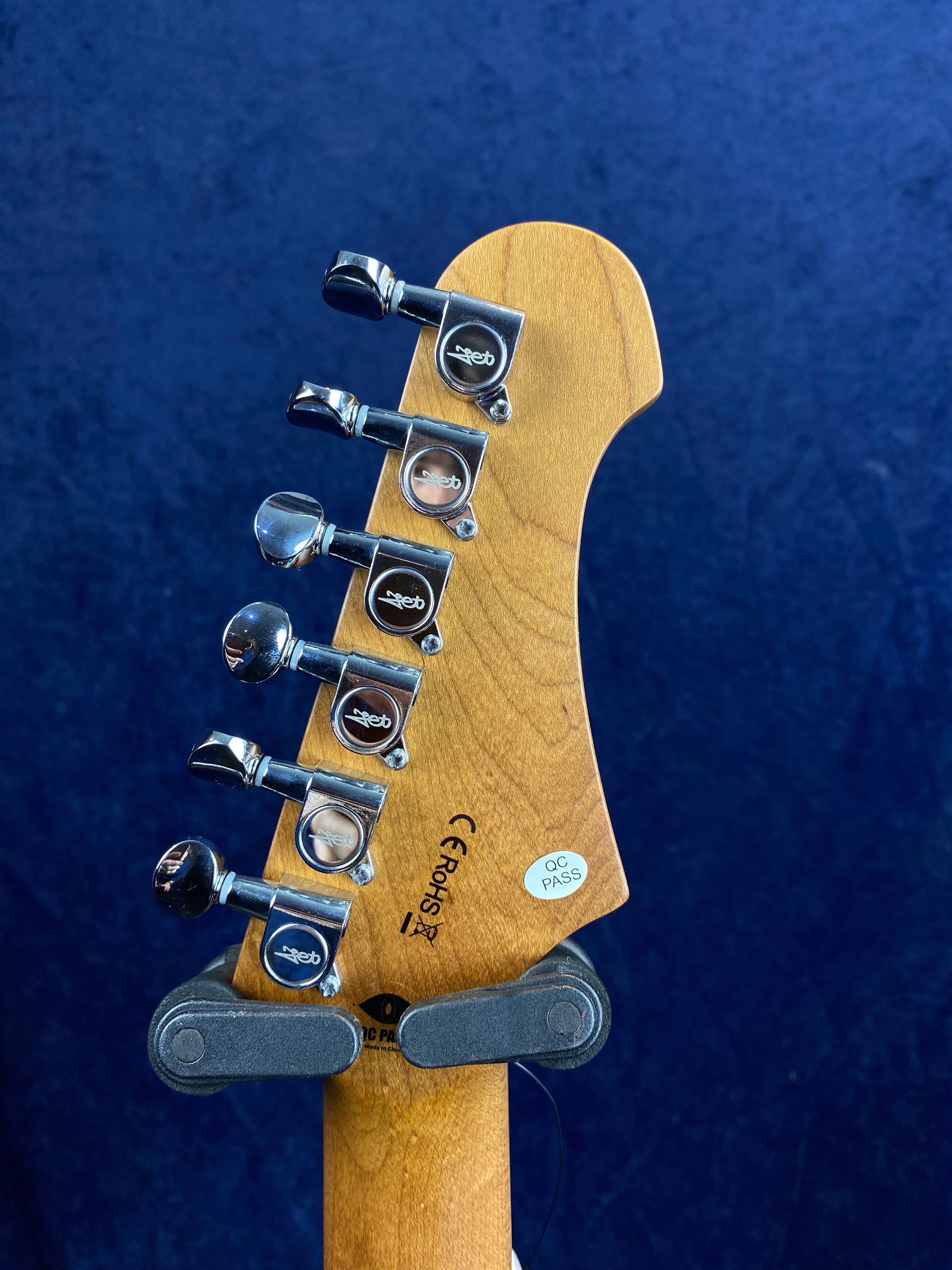 Six chrome tuning pegs on a light wood electric guitar headstock, with "QC PASS" and certification logos visible against a dark blue background.