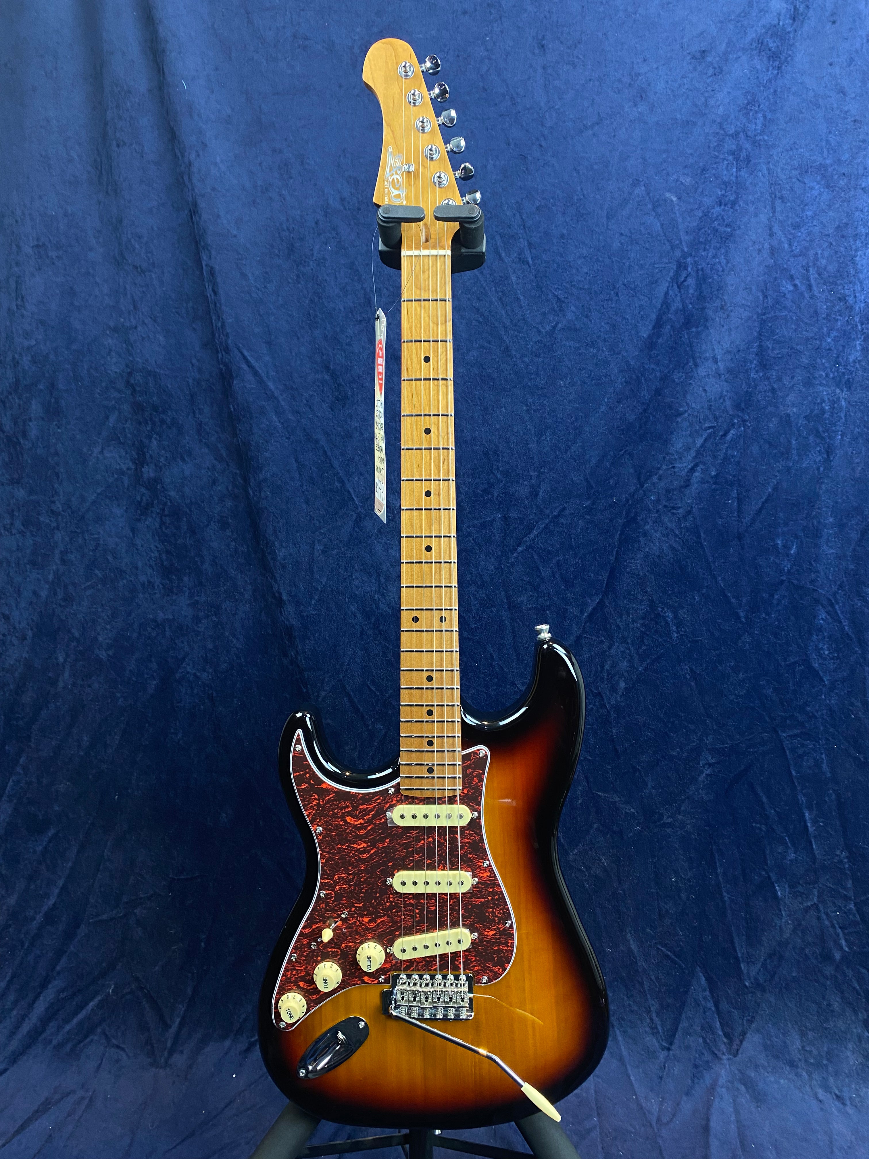 Sunburst-finish electric guitar with a maple neck, three single-coil pickups, tortoiseshell pickguard, and cream-colored knobs, displayed on a stand.