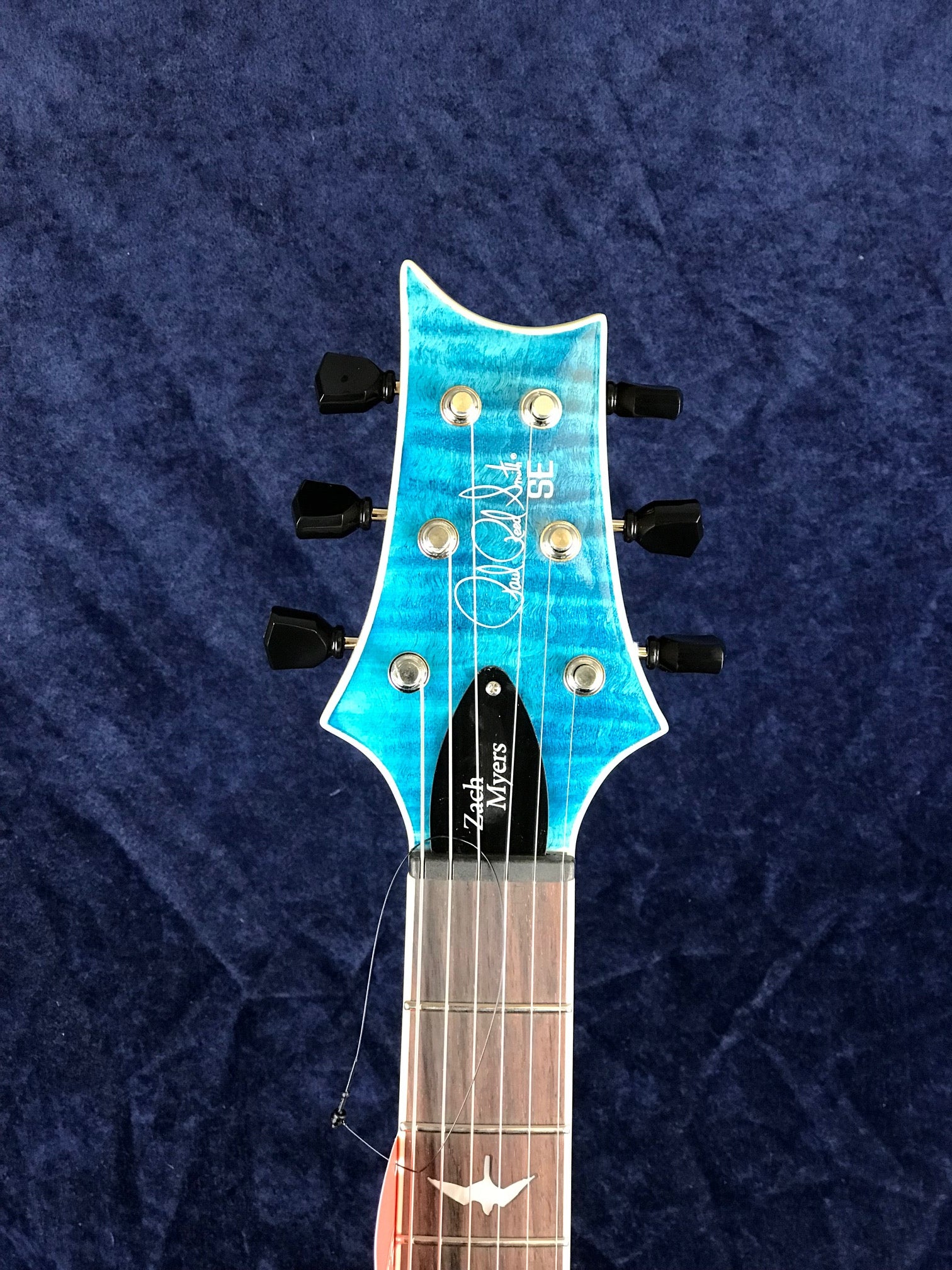 Electric guitar headstock with a blue finish, six black tuning pegs, Zach Myers signature, and white inlay bird on the rosewood fretboard.