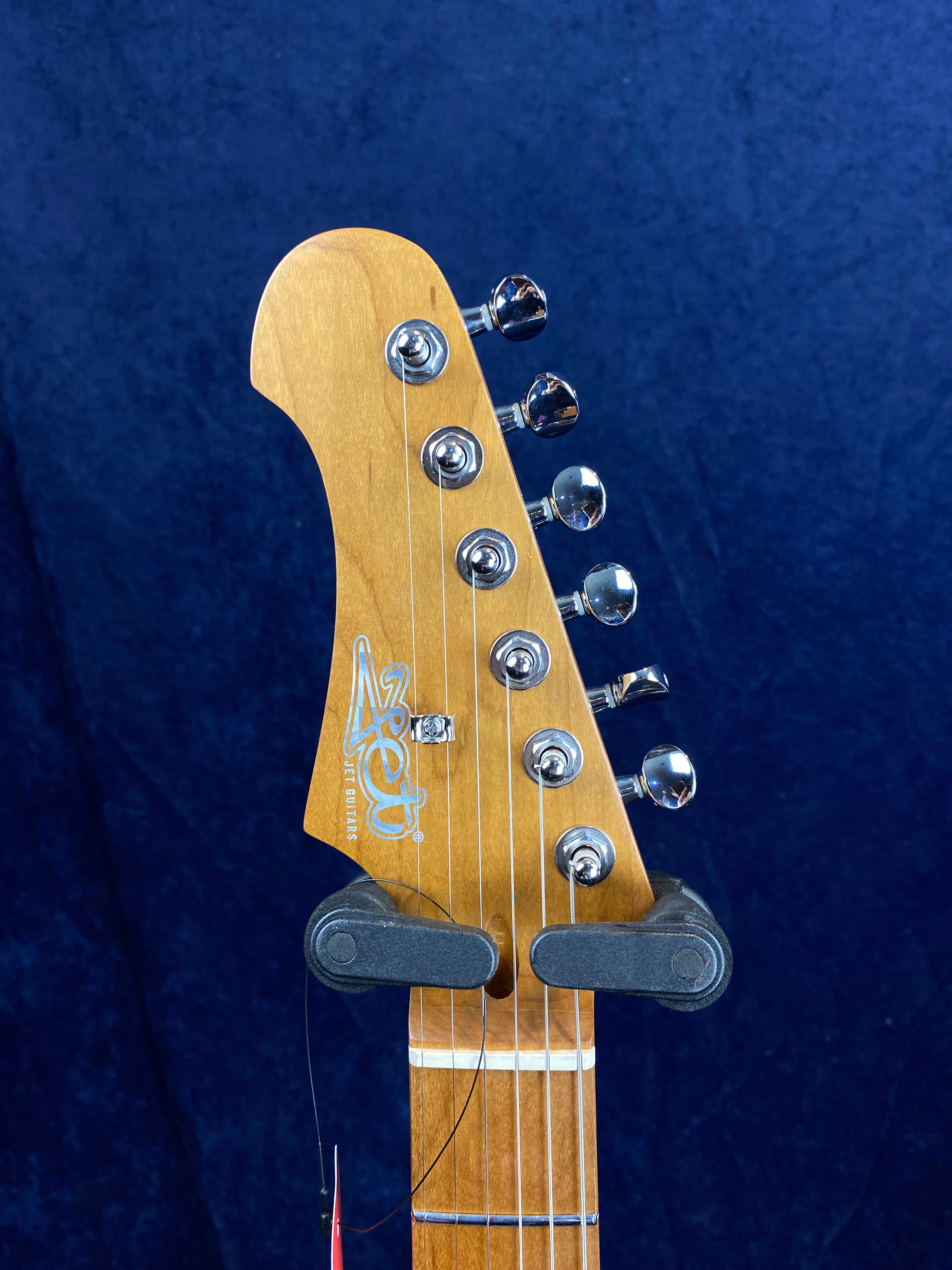 Light brown Jet Guitars electric guitar headstock with six chrome tuning pegs against a dark blue background.