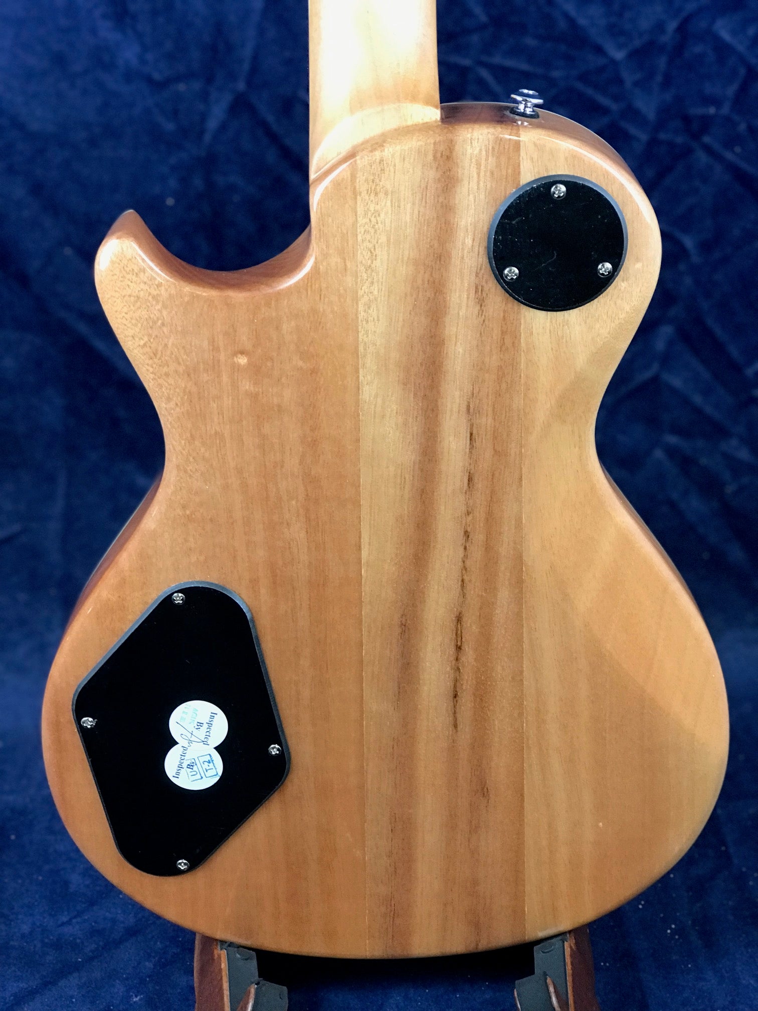Back of a natural wood electric guitar body showing two black plastic compartment covers with screws, one round and one irregularly shaped.