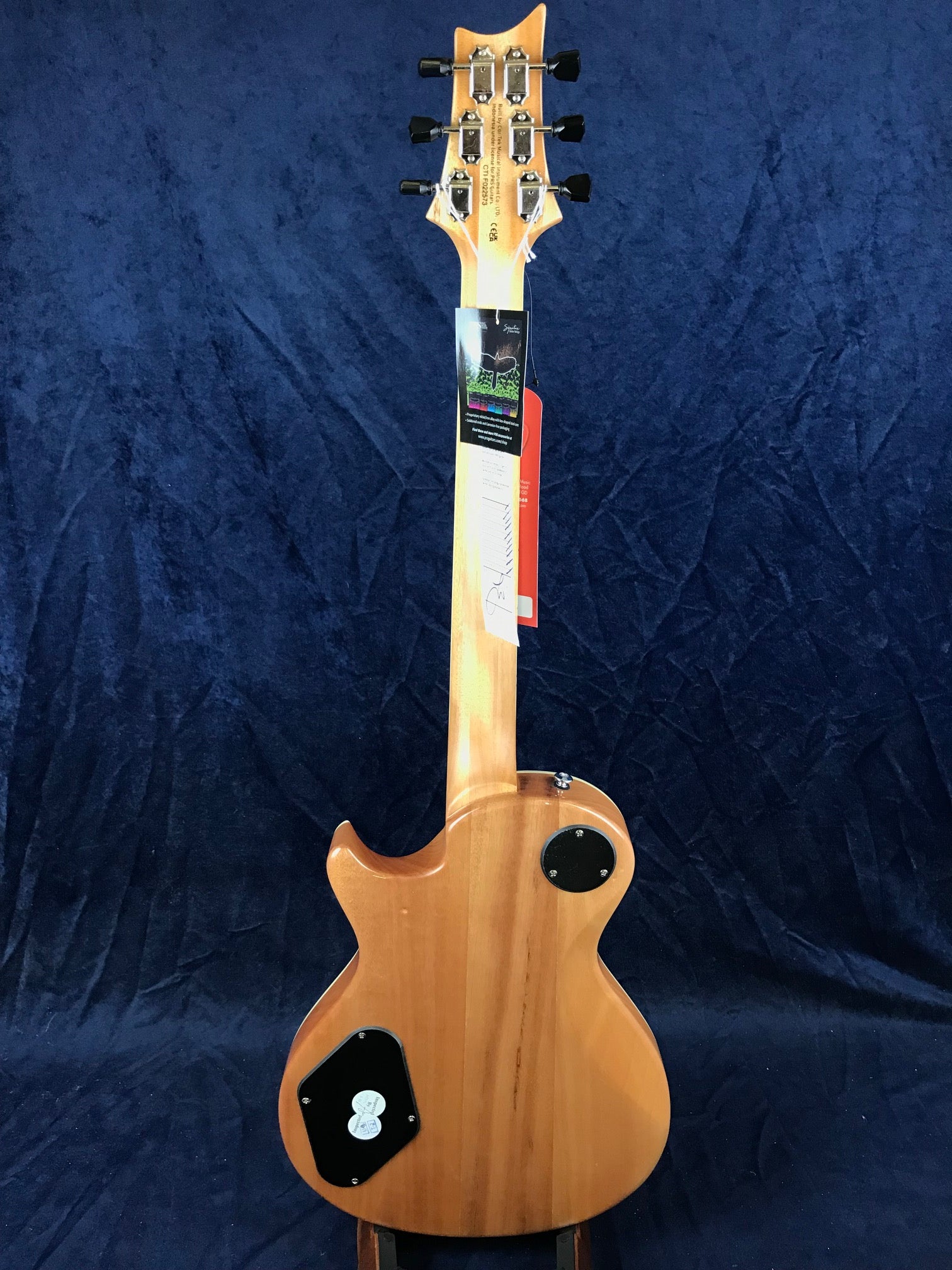 Natural wood finish electric guitar seen from the back, showing tuning pegs, control cavity covers, and factory tags hanging from the neck.