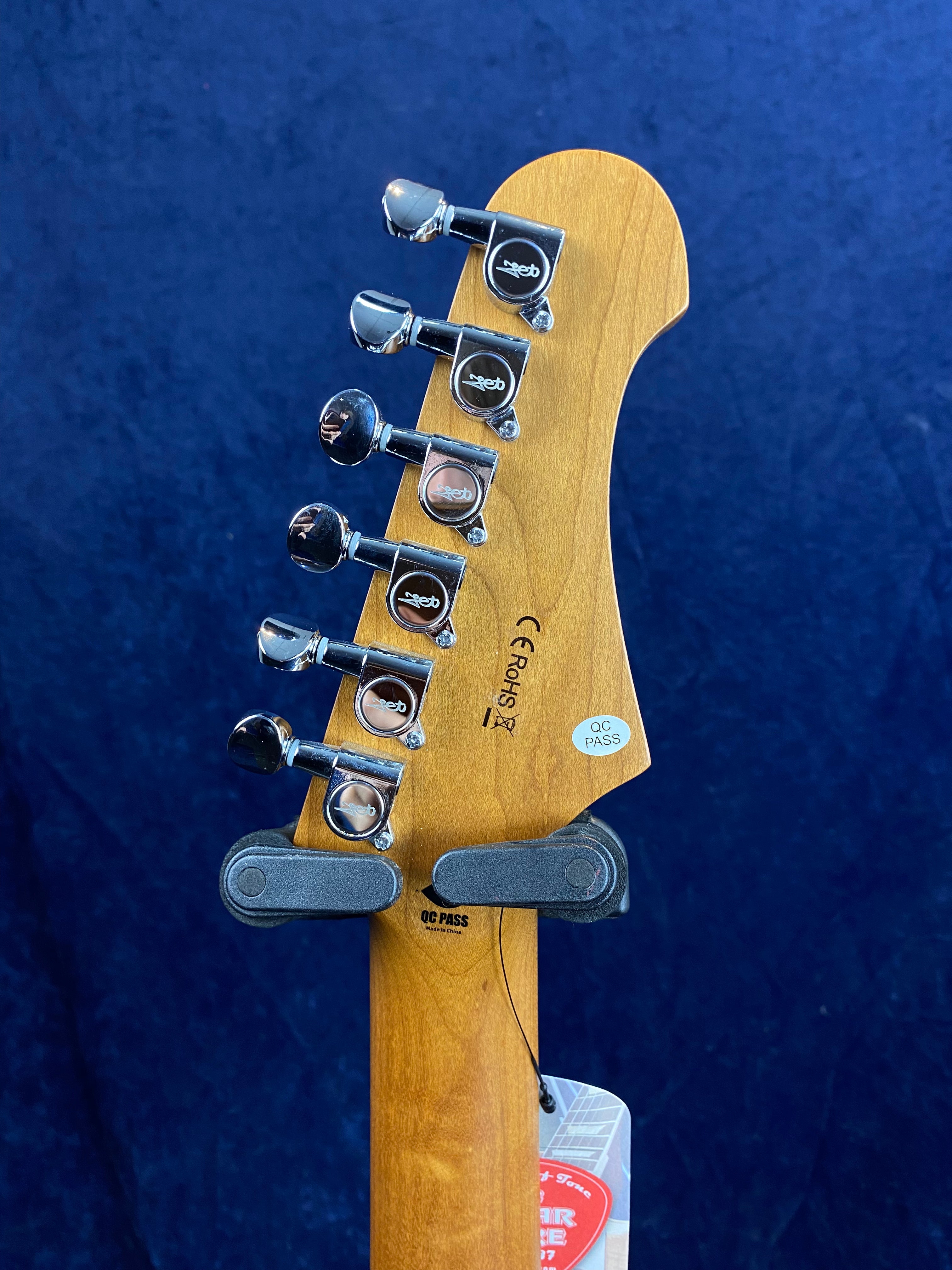 Six chrome tuning pegs on the headstock of a wooden electric guitar neck, with QC pass stickers and a hang tag visible.
