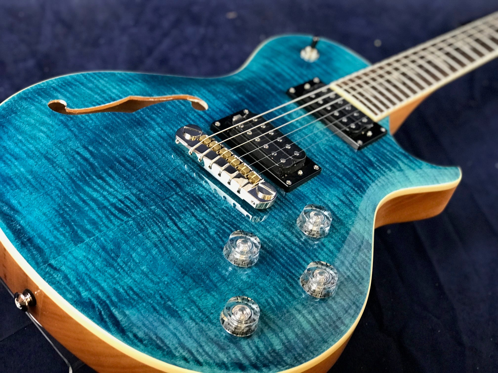 Electric guitar with a glossy blue flamed finish, f-hole cutout, two humbucker pickups, four clear control knobs, and a wood-trimmed edge.