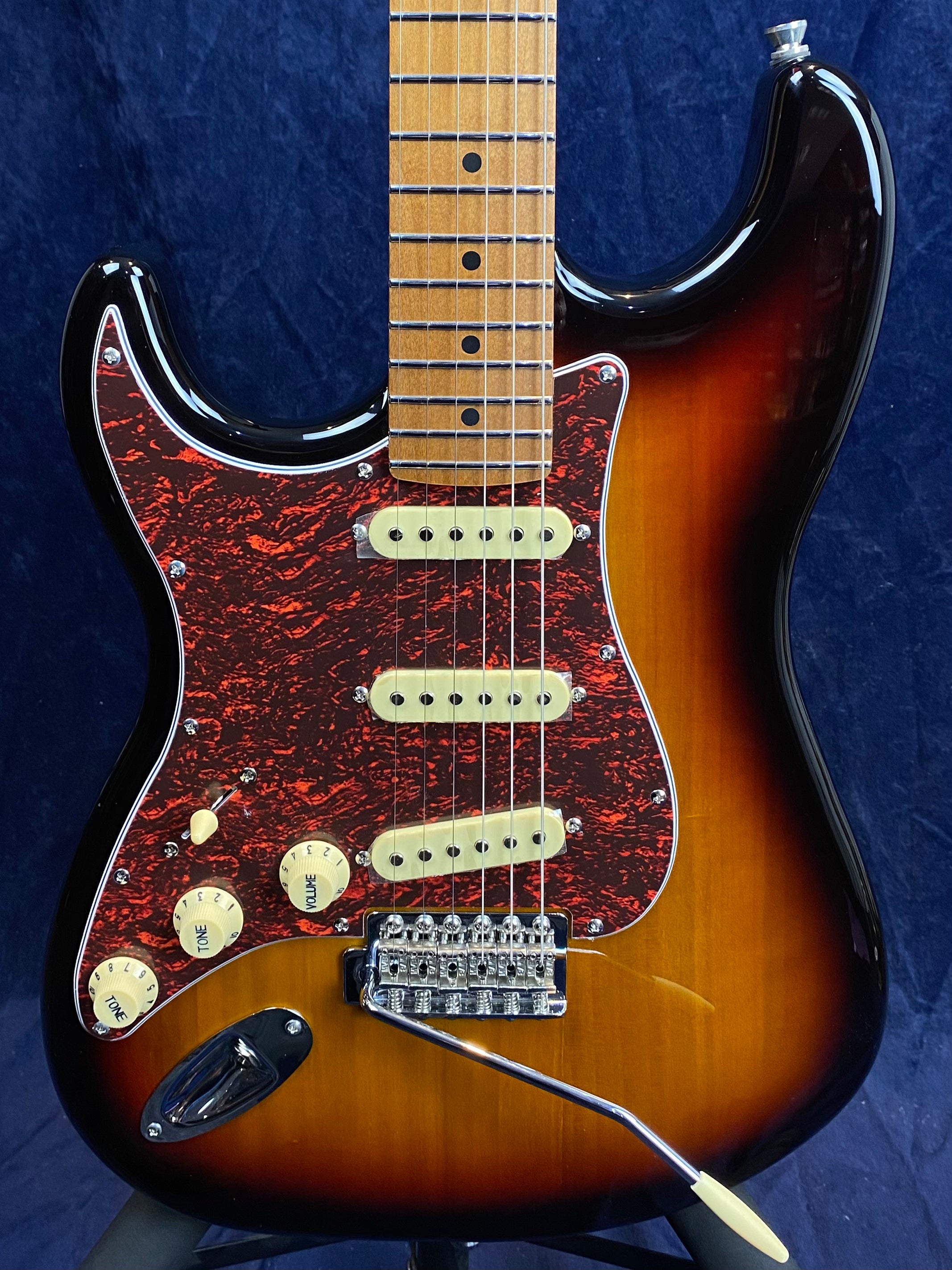 Electric guitar with a sunburst finish, red and black pickguard, three single-coil pickups, and a maple fretboard.