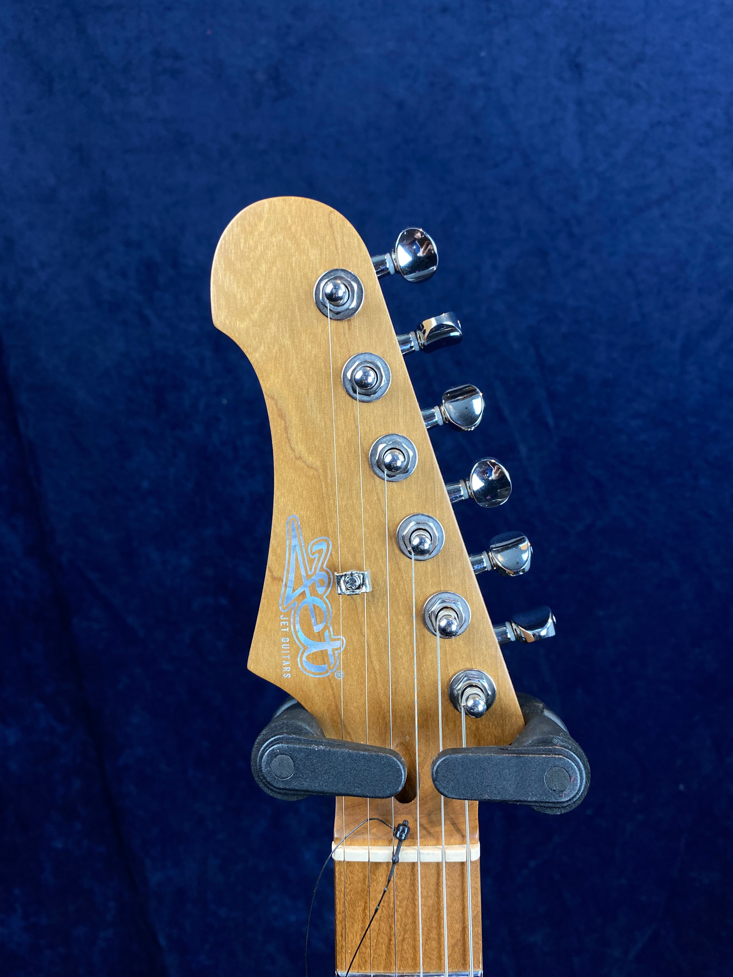 A light wood electric guitar headstock with six chrome tuning pegs, Jet Guitars logo in silver, and a guitar holder gripping the neck.