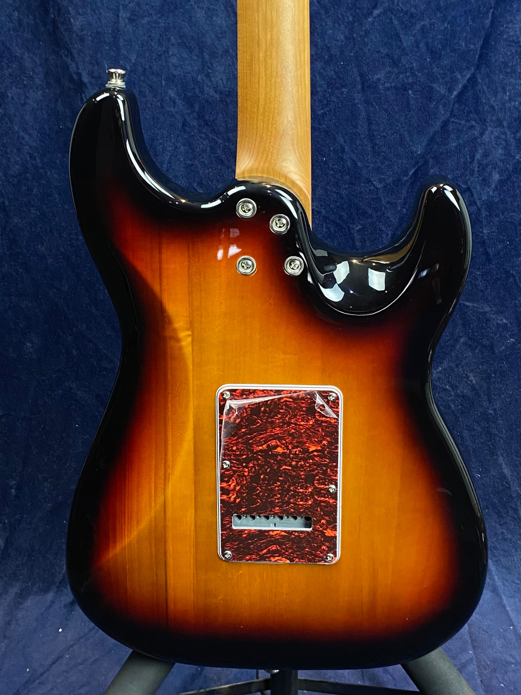 Sunburst-finish electric guitar back, tortoiseshell-patterned cover plate, three neck bolts visible, natural wood neck, dark blue background.