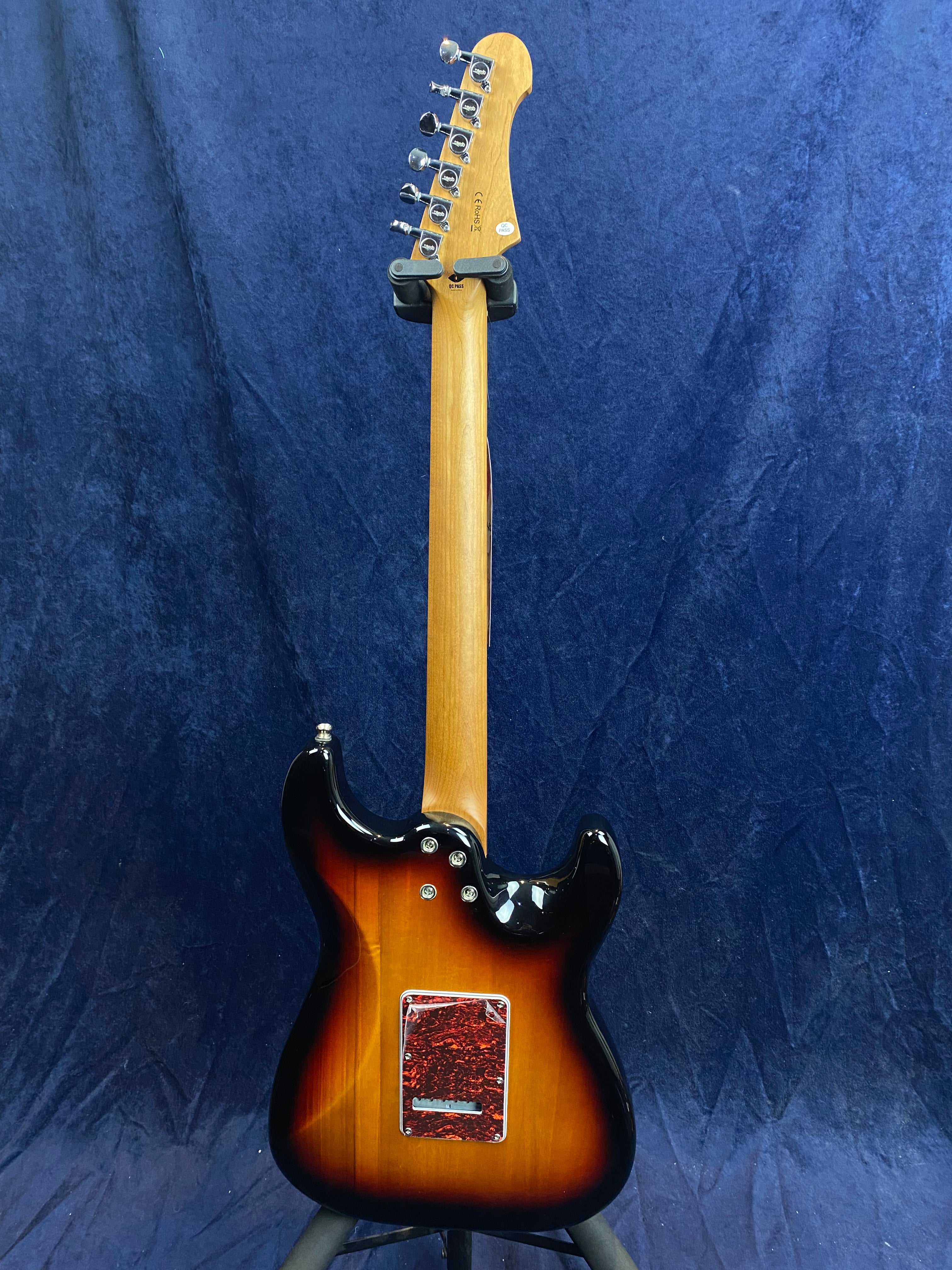Sunburst electric guitar shown from the back with a natural wood neck and red patterned cover plate, standing against a dark blue fabric background.
