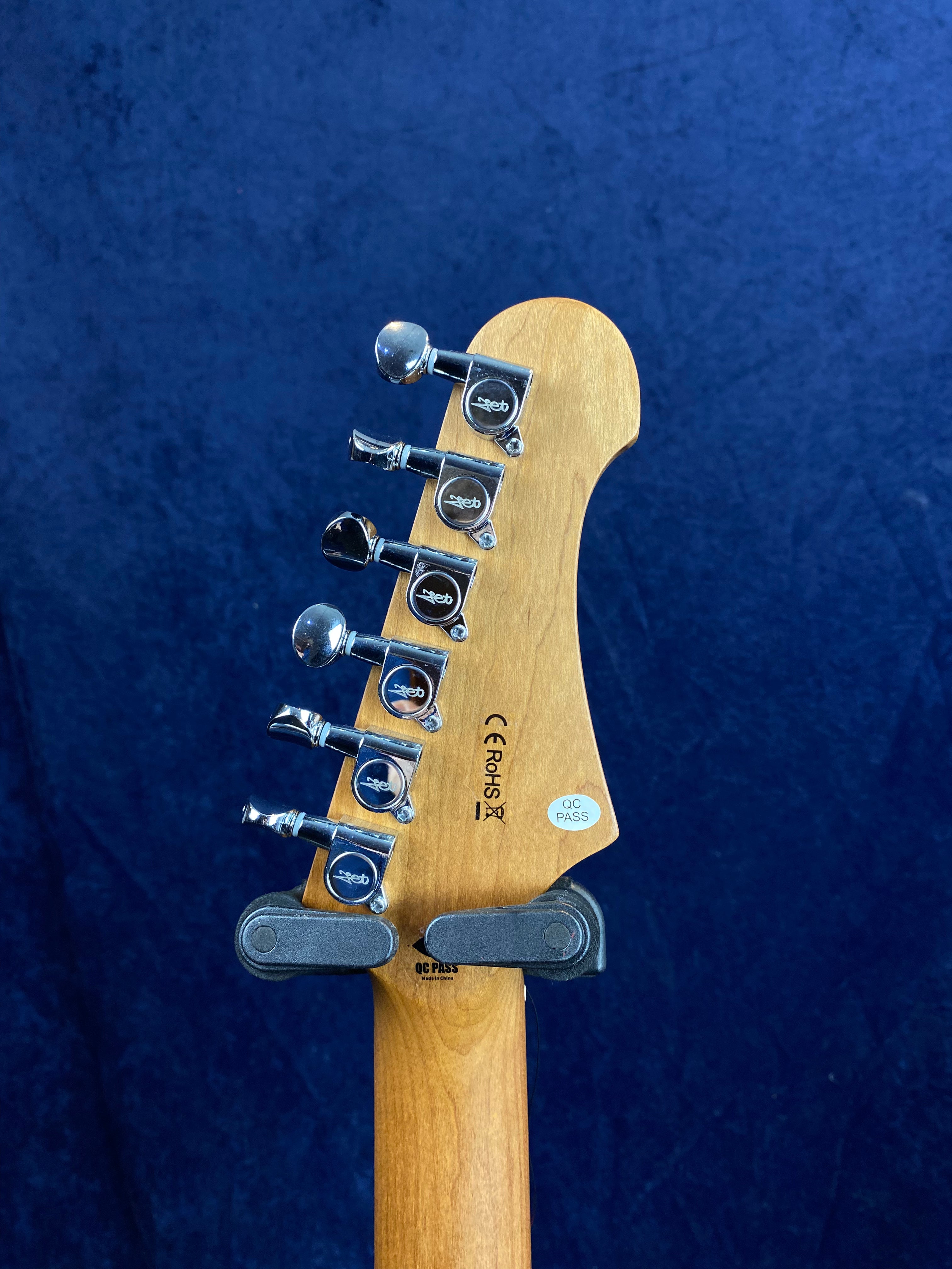 Six metal tuning pegs line the headstock of this electric guitar, with certification stickers and a wooden finish visible against a dark blue background.