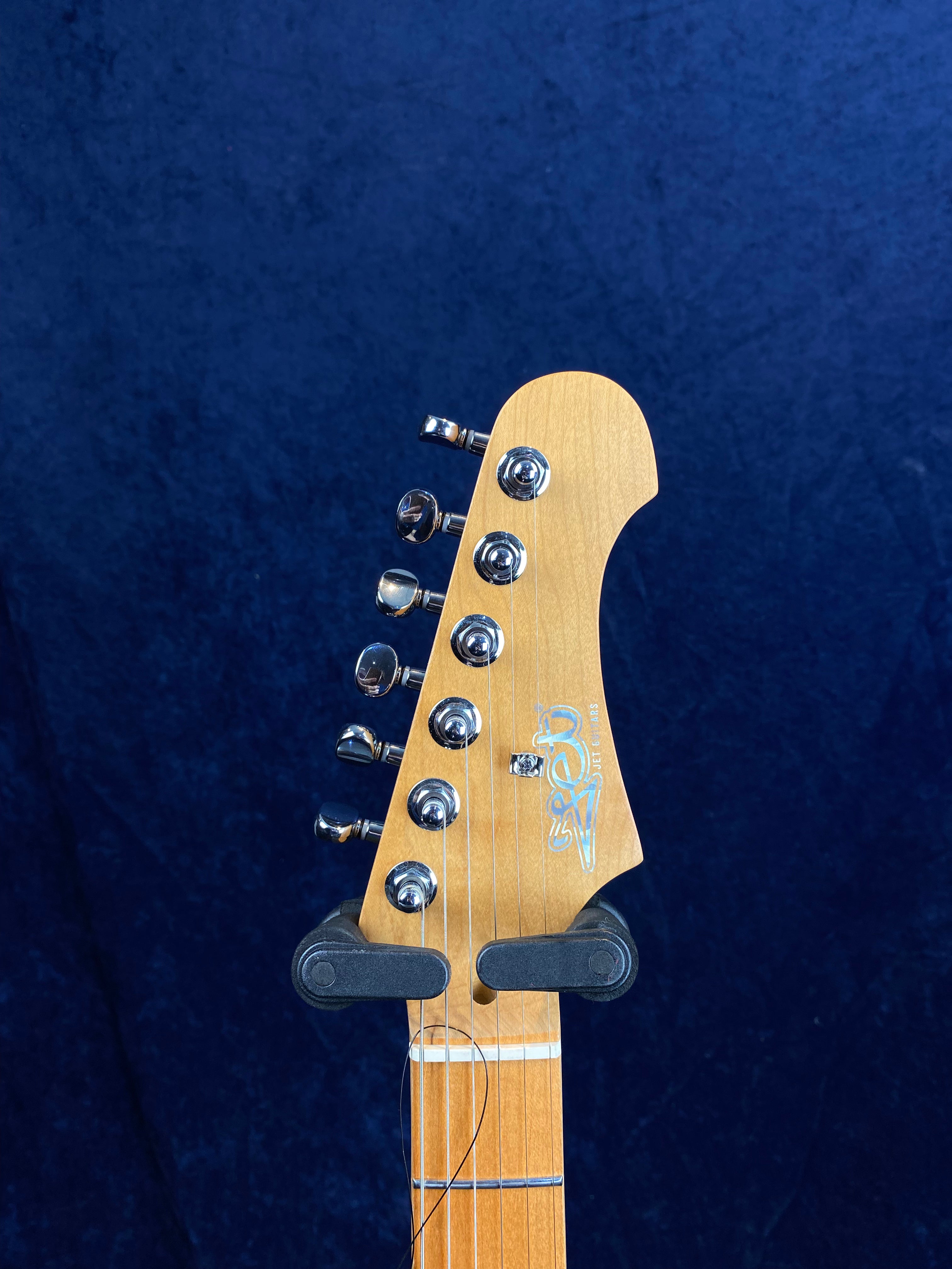 Light wood electric guitar headstock with six chrome tuning pegs, logo, and strings against a solid dark blue background.